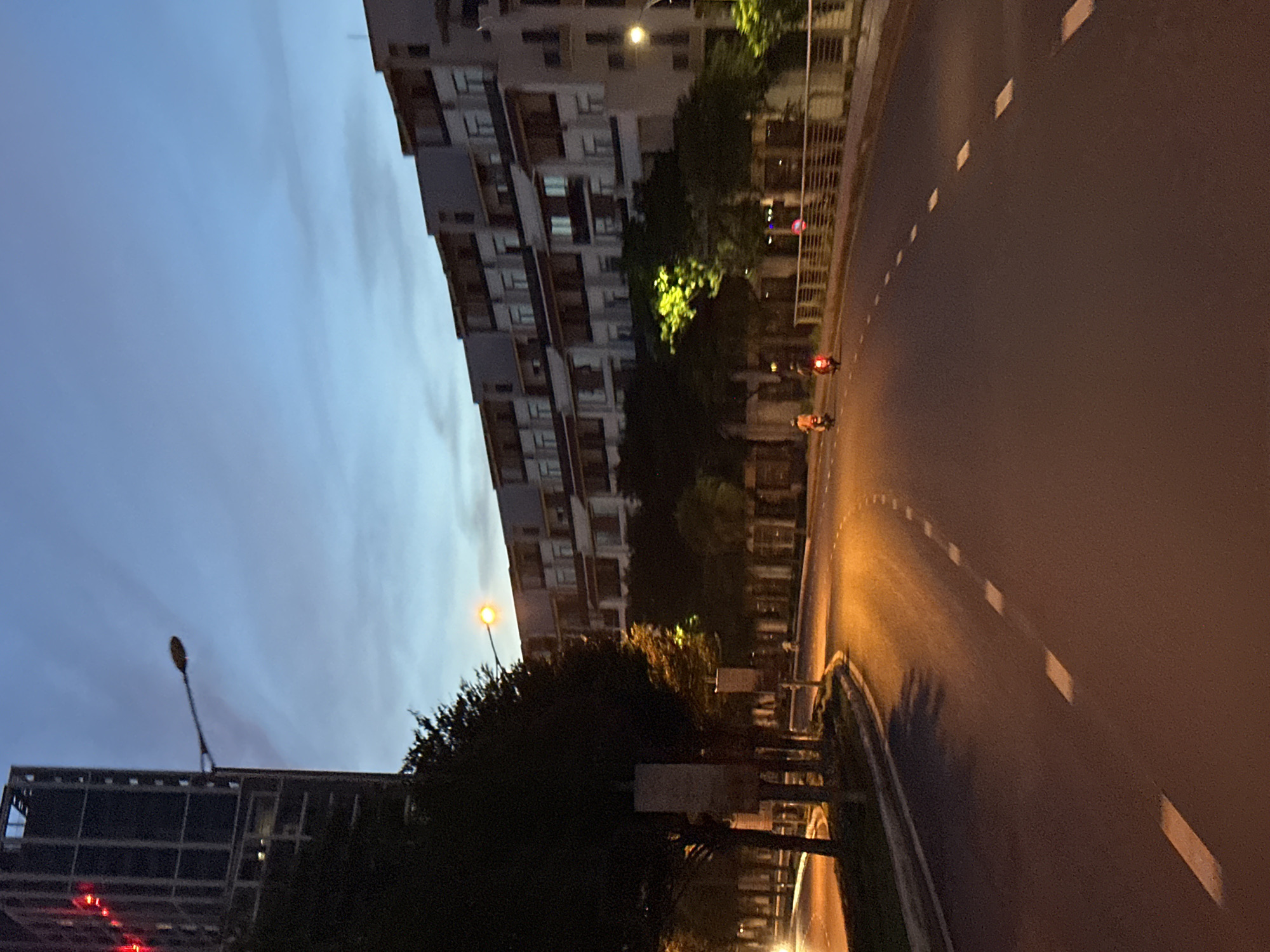 The image depicts a wide asphalt road in the city of Da Nang, Vietnam, during twilight. A row of modern, multi-story buildings with balconies lines the right side of the road, illuminated by streetlights and interior lighting. Trees with lush green foliage are visible along the left side of the road, casting shadows on the asphalt. Two individuals on motorcycles are traveling on the road, their headlights and taillights creating streaks of light. The sky is a gradient of deep blue and lighter blue, suggesting the end of daylight. Streetlights illuminate parts of the road and the surrounding vegetation. The road features white dashed lines for lane separation. The overall scene conveys a sense of urban transit and the transition from day to night.
