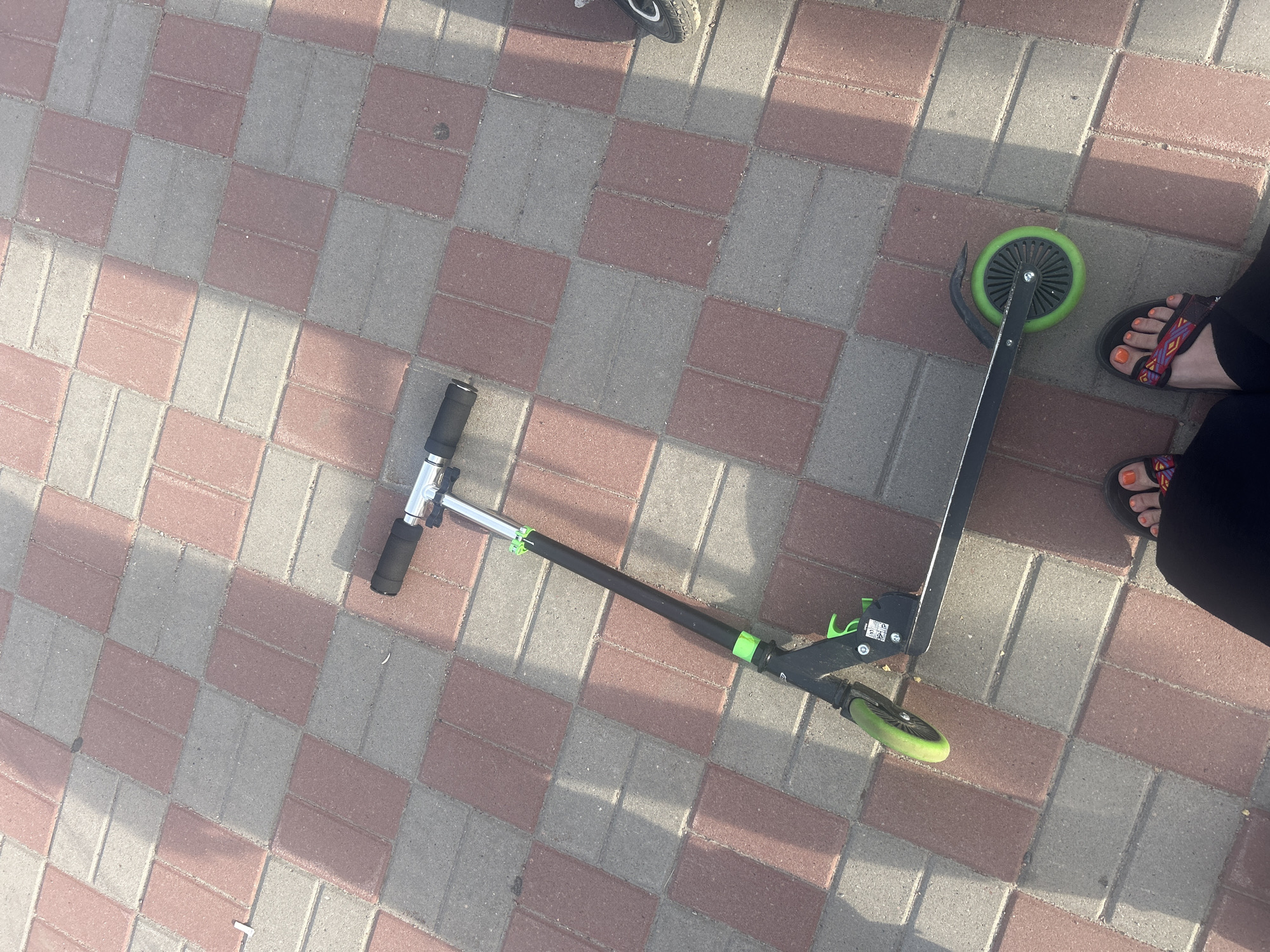 A black and green kick scooter is lying on a paved walkway with red and gray rectangular bricks arranged in a checkerboard pattern. The scooter is positioned diagonally across the frame. The scooter has black hand grips, a silver steering column with green adjustment clamps, and a black frame with green accents. It has two green wheels, one at the front and one at the rear. Shadows from an unseen object, possibly a tree or building, are cast across the walkway. In the lower part of the image, a person's feet are visible, wearing black pants and patterned sandals with orange nail polish. The scene appears to be outdoors in daylight.