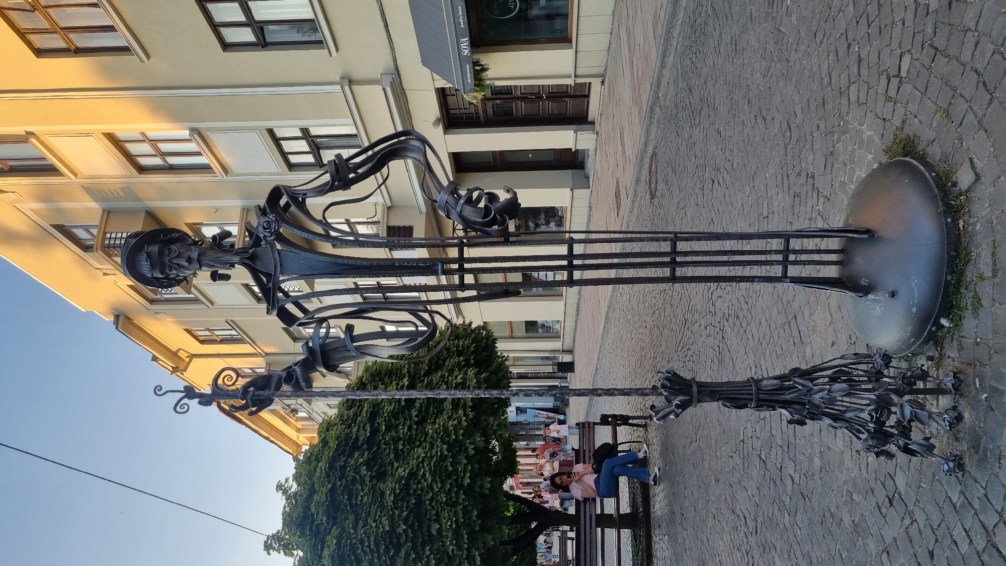 In Chernivtsi, Ukraine, a metal sculpture of a man stands on a cobblestone street in front of a building with cream-colored walls and brown-framed windows. The sculpture is tall and slender, made of wrought iron, and depicts a figure with a hat, glasses, a mustache, and a beard, holding a staff topped with decorative elements in its left hand and what appears to be a bundle of metal flowers at its base. A woman sits on a park bench to the left of the sculpture, looking at her phone. Behind her, people are walking and a large, leafy tree provides shade. The street is paved with cobblestones, and buildings line both sides of the street. The sunlight casts shadows, indicating it is late afternoon.