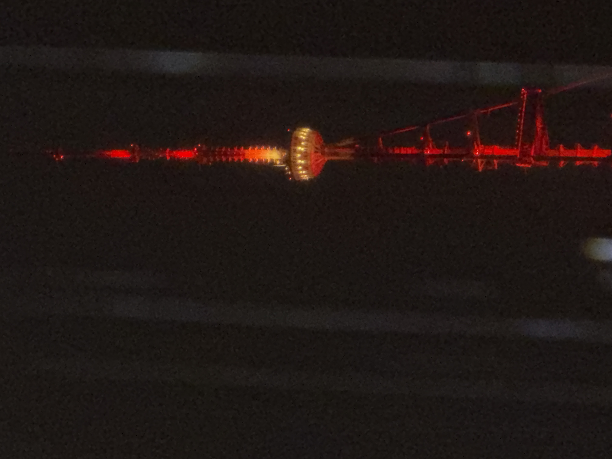 The image shows a night view of illuminated structures reflected in water. The central focus is a tower-like structure with a distinct, brightly lit central sphere, likely a broadcasting or communications tower. To the right of the tower, a bridge structure with red lights along its span is visible. The dark background indicates it is nighttime, and the reflections suggest the presence of a body of water like a river or lake. Based on the illuminated structures and the description, the location appears to be Tbilisi, Georgia. The lights are vivid and create a sharp contrast against the darkness.