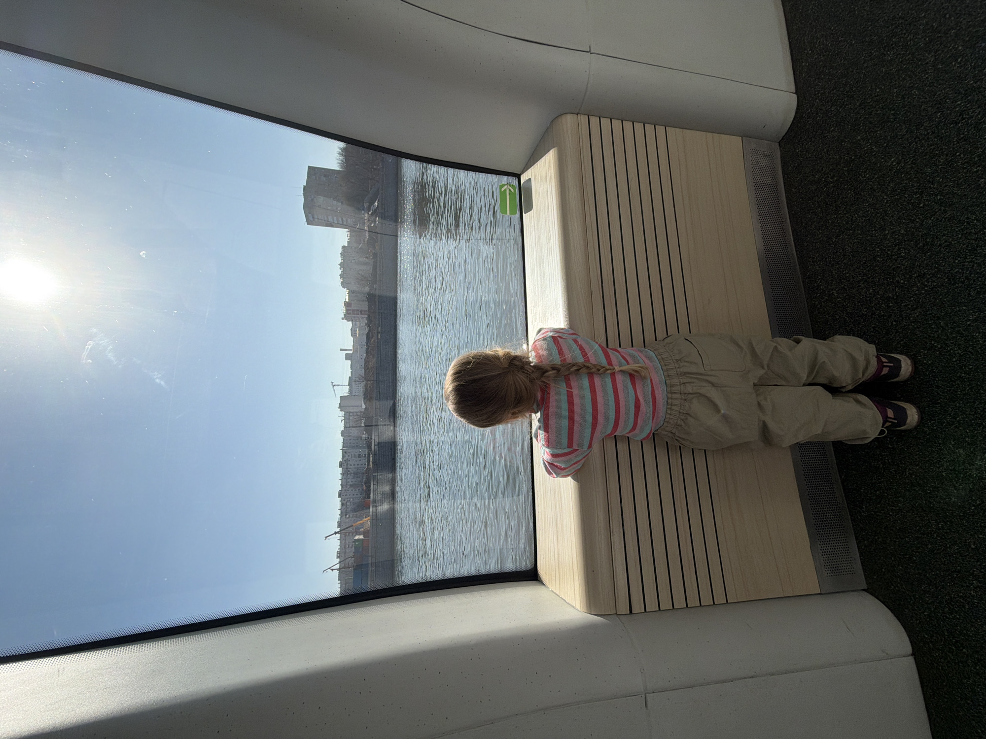 A lone child, viewed from behind, stands inside a modern electric river tram, gazing out a large panoramic window onto the Moskva River in Moscow, Russia. The child, a young girl with a single blond braid, wears a horizontally striped long-sleeve shirt (pink, blue, white, and light grey), tan pants, and dark sneakers with maroon accents. She leans forward, resting her forearms on a light-colored wooden console with horizontal slat detailing, which serves as a window sill. The interior features curved white walls and a dark green speckled floor. A small green sticker with a white right-pointing arrow is visible on the windowpane to the right.
Through the window, under a clear, bright blue sky, the sun is high in the upper right, causing a lens flare effect. The wide Moskva River extends across the view, its surface exhibiting gentle ripples and reflections. On the opposite bank, a prominent concrete embankment wall fronts a line of bare trees, beyond which stands a mix of urban buildings. These include multi-story residential blocks and a distinctive, angular-topped building further to the right. Several large construction cranes, orange and white, are visible on the far bank, indicating active development or industrial operations.