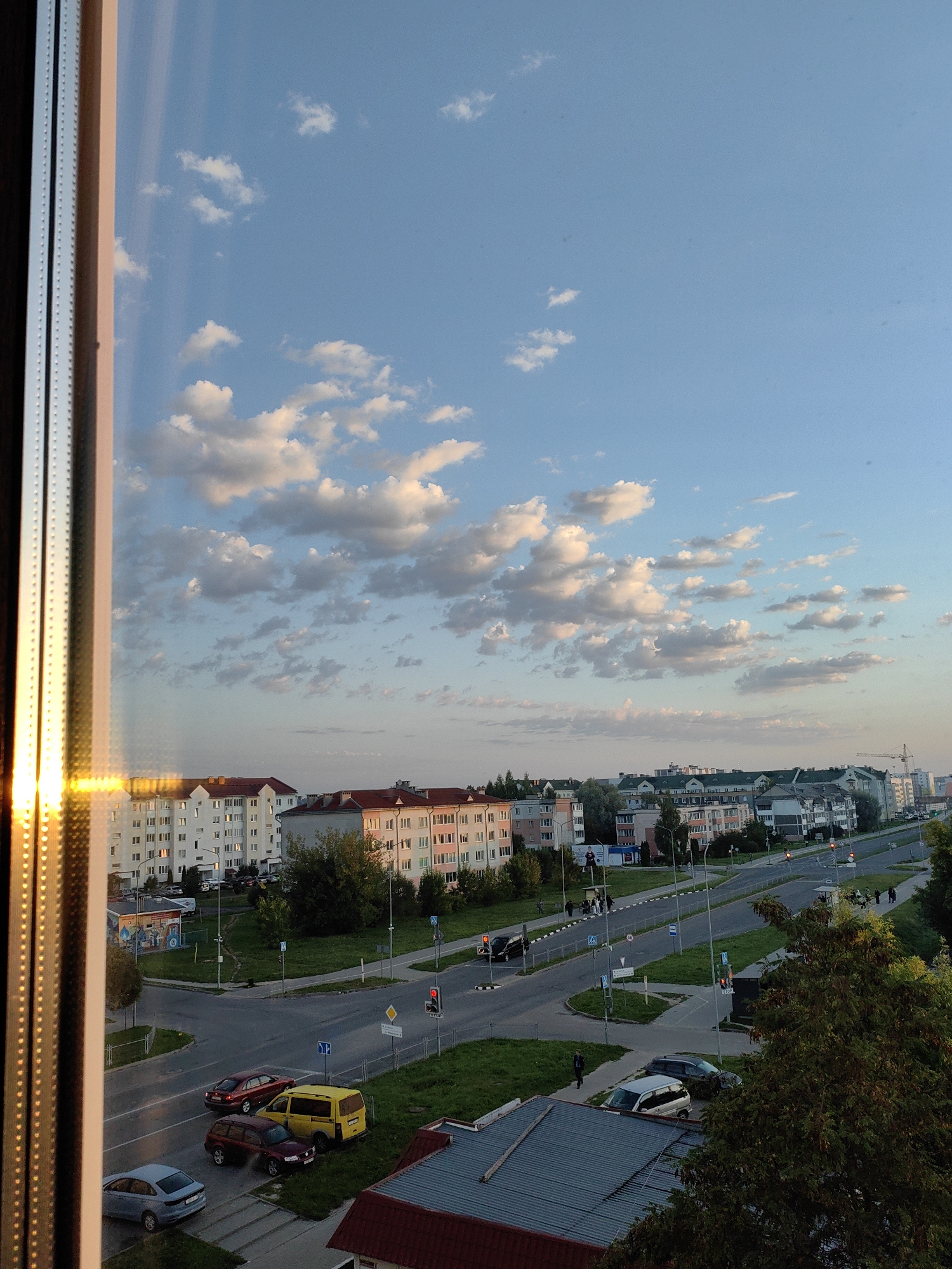 This is an aerial view of a city intersection in Fanipal, Belarus, at dusk. The sky is a soft blue with scattered white clouds tinged with pink from the setting sun. In the foreground, a window frame with reflective golden light runs along the left side of the image. The scene below shows a multi-lane road with several cars, including a red sedan, a burgundy station wagon, a yellow minivan, and a light blue sedan. Pedestrians are visible on sidewalks and at a bus stop. Buildings lining the street are multi-story apartment complexes with pitched roofs, characteristic of Eastern European architecture. A construction crane is visible in the distance. The environment appears to be a typical urban residential area with green spaces along the road.
