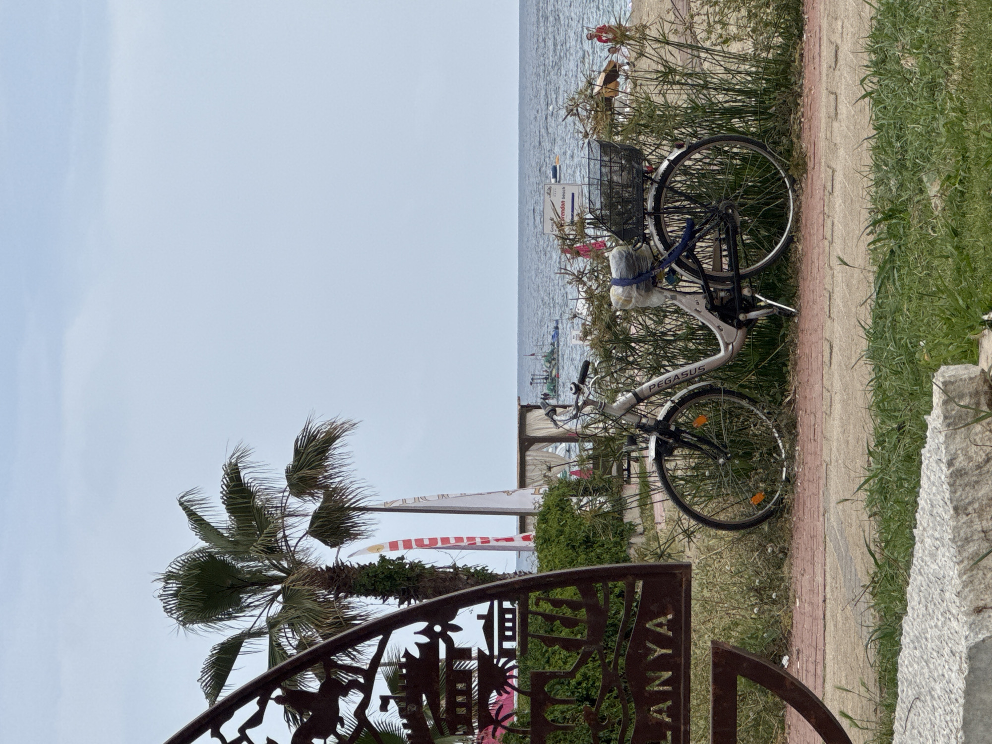 A landscape photo captures a beach scene in Alanya, Turkey. The beach is visible in the background with people and some boats. In the foreground, there is a bicycle, a palm tree, and a metal gate. The word "Alanya" is written on the gate.