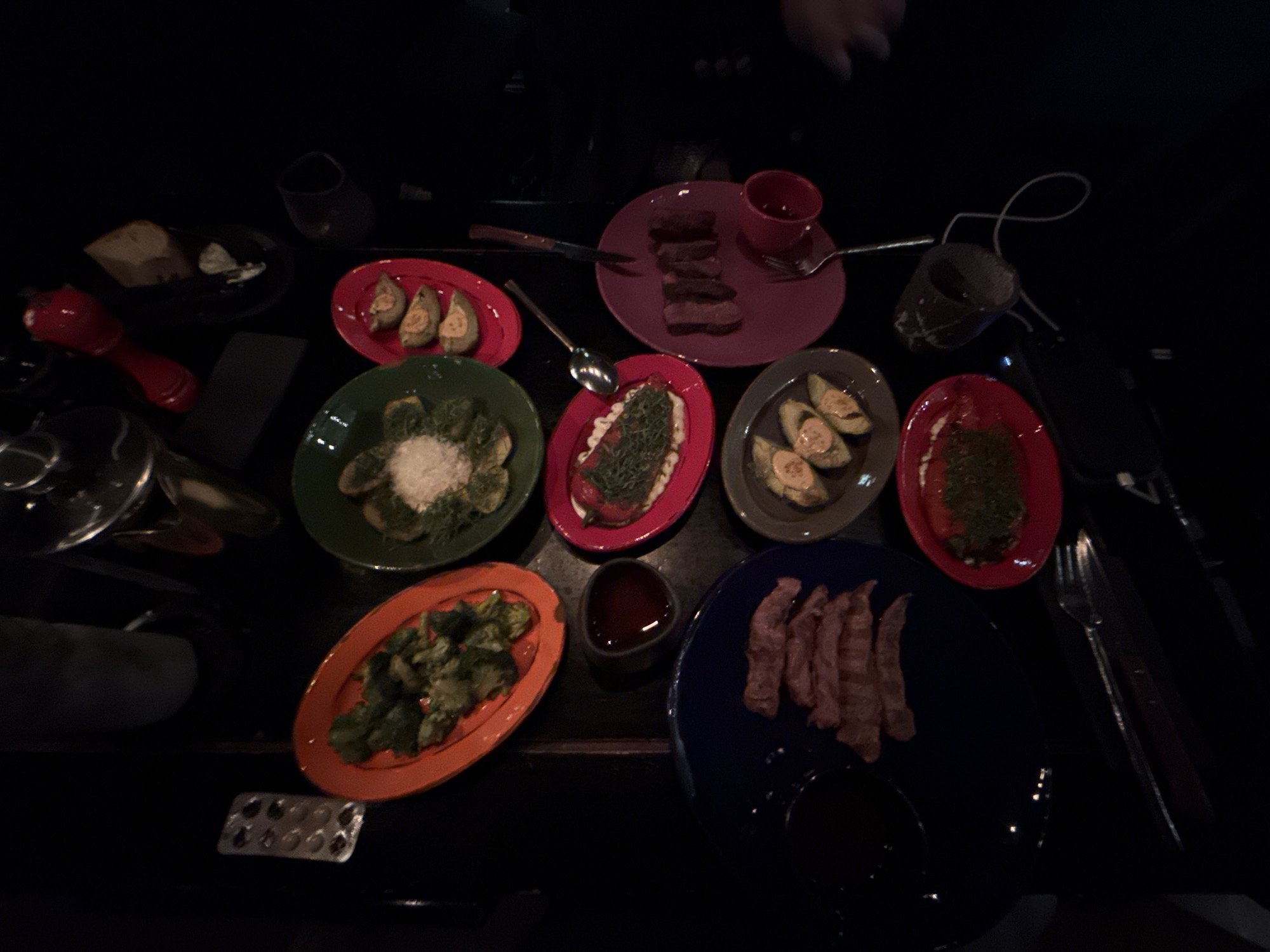 The image depicts a dark, low-lit setting, likely a restaurant table in Moscow, Russia, given the likely Eastern European cuisine, featuring an array of small dishes of food served on colorful plates. From left to right, the dishes include bread with some sort of spread, a dish of what appears to be stuffed artichokes, followed by a green salad with parmesan cheese on top, a red plate with salmon salad, a grey plate with a type of stuffed avocado, then a red plate with another green salad and last is a blue plate with slices of meat. Small dishes of sauces accompany the food as well as an empty blister pack of medicine. Cutlery and drinking glasses are also visible. The overall atmosphere is sophisticated.