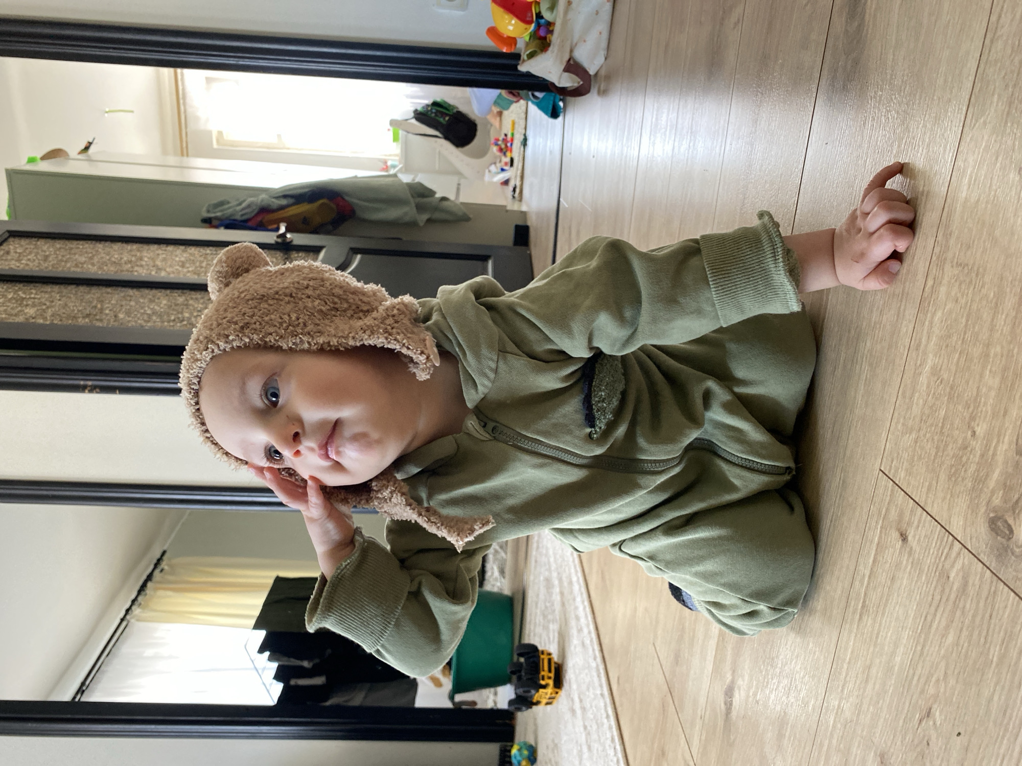 A young child wearing a brown bear-eared hat and an olive-green jumpsuit lies on a light-brown, wood-patterned floor. The child rests on their side, with one hand touching their head and the other resting on the floor. Reflected in a dark-framed mirror behind the child is a room with white walls, a dresser, and other items suggesting a domestic environment. A small yellow and black toy truck sits on a light-colored rug near the child. Additional toys are scattered in the background, beyond the mirror's reflection. The scene appears to be indoors, likely in a residence. There is no information about the location in the city of Chaikovsky, Russia.