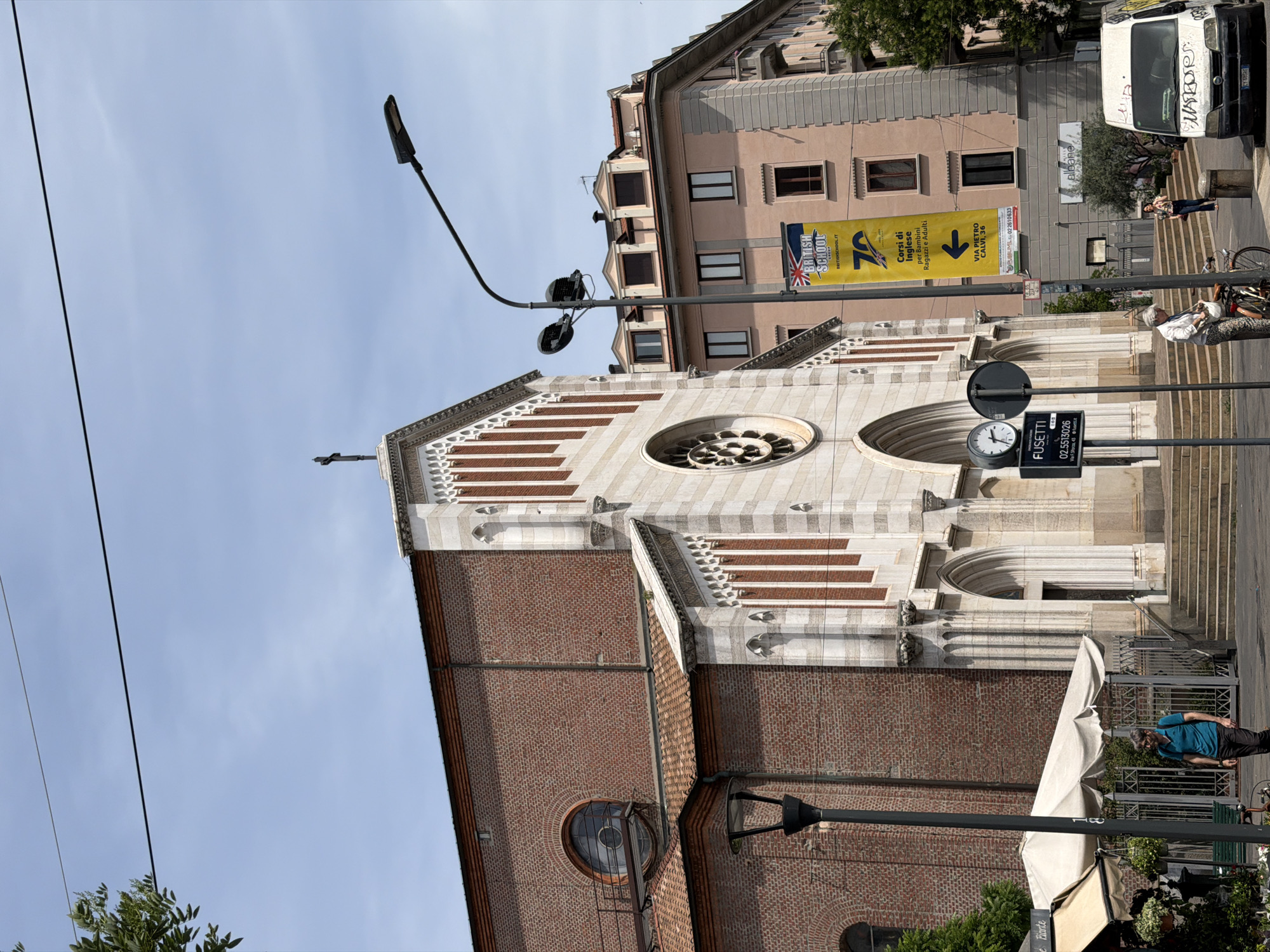 This is an eye-level, medium shot of a church facade on a sunny day in Milan, Italy. The church is constructed of red brick with white stone accents. The facade features a large rose window and multiple arched doorways. A banner for the "British School" hangs from a lamppost in front of a pink building behind the church. Several people are visible, including a man walking on the sidewalk, a woman with a bicycle, and a woman standing near a parked van. The environment includes a street, sidewalks, trees, and buildings. The sky is clear and blue.