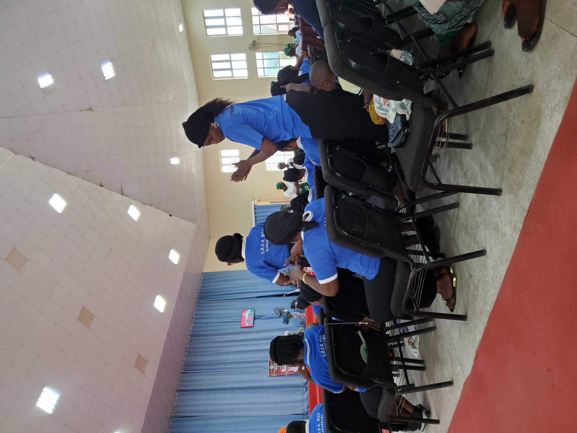 The image shows a gathering of people, primarily women, in what appears to be a large indoor space, possibly a church or community hall. The women are mostly wearing blue shirts with a logo or text visible, possibly indicating a uniform or group affiliation. A portion of the text on the shirts says "S.R.D.K RCC MARARABA." They are seated in black folding chairs arranged in rows. Some are engaged in actions such as clapping or looking down, seemingly focused on something happening in front of them. The room has a high ceiling with integrated lighting and windows providing natural light. The flooring is partially covered with what looks like a red carpet. The setting is in Mararaba, Nigeria, as indicated by the text on the shirts.
