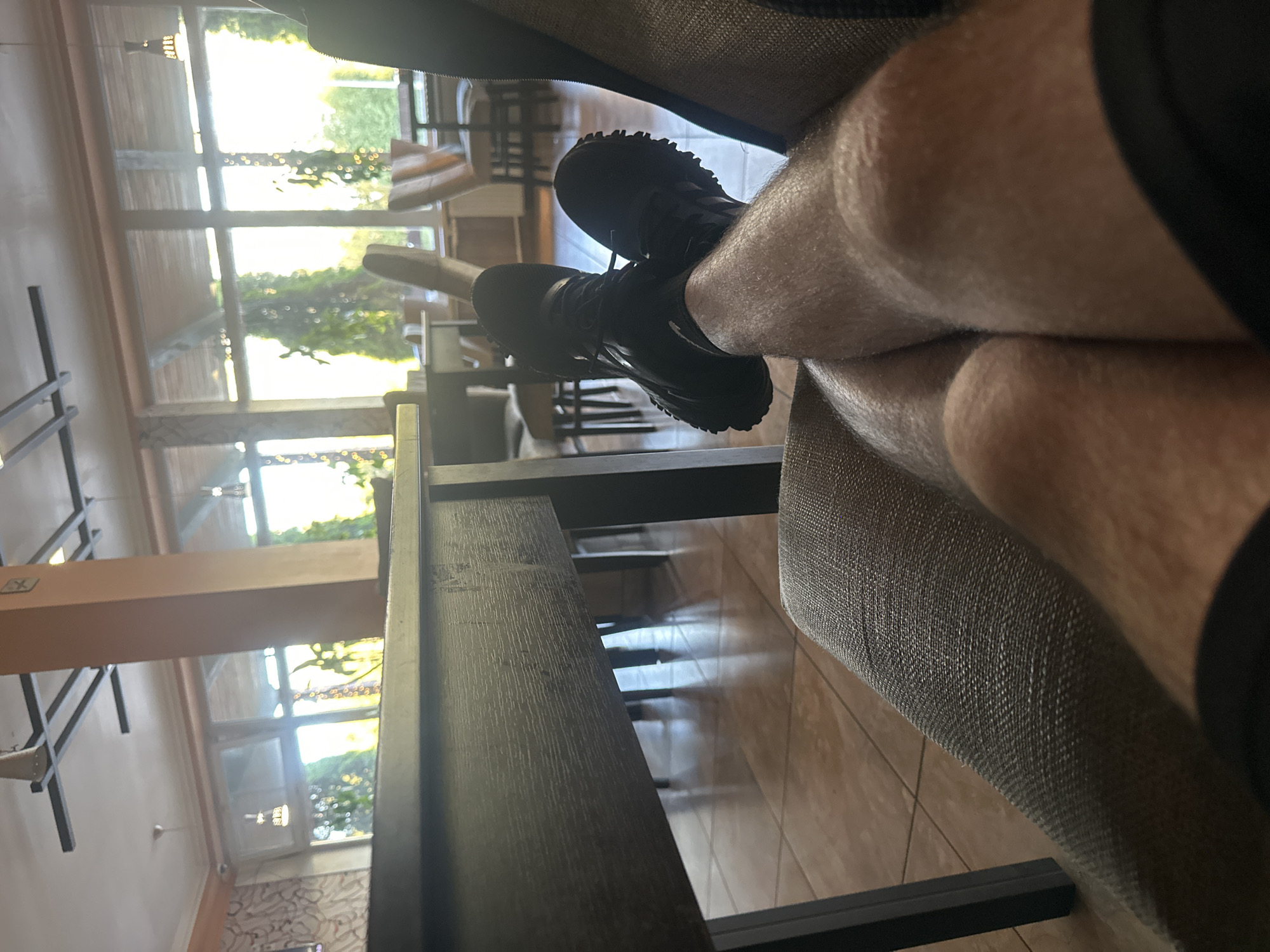 A first-person perspective shot captures a person relaxing indoors, with their legs crossed and resting on a textured chair. The person is wearing black athletic shoes and shorts, with visible leg hair. The setting appears to be a cafe or restaurant, suggested by the presence of tables and chairs visible through a large window. Outside the window, lush greenery and trees are visible, bathed in bright sunlight. The indoor environment features a large, dark wooden table in the foreground and light-colored tiled flooring. The decor includes a tall, light-brown pillar and subtle wall patterns. The overall impression is one of a tranquil moment in a well-lit, pleasant establishment, possibly in a suburban or rural setting, given the prominent natural elements outside. The location is identified as Zapolyev, Russia.