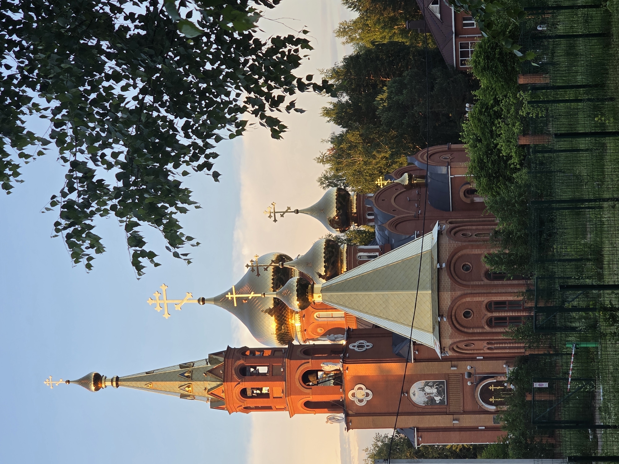 The image shows the Holy Ascension Cathedral, a prominent landmark in the city of Chaikovsky, Russia. The cathedral is a large, brick structure with multiple golden domes, characteristic of Russian Orthodox architecture. A tall bell tower with a pointed roof stands on the left side of the building. The sun is setting, casting a warm, golden light on the cathedral and its surroundings. Lush green trees and foliage surround the cathedral, and a metal fence is visible in the foreground. The sky is a soft blue with hints of orange and pink from the sunset. There are no people visible in the image. The overall scene is peaceful and serene, highlighting the beauty of the cathedral and its natural setting.