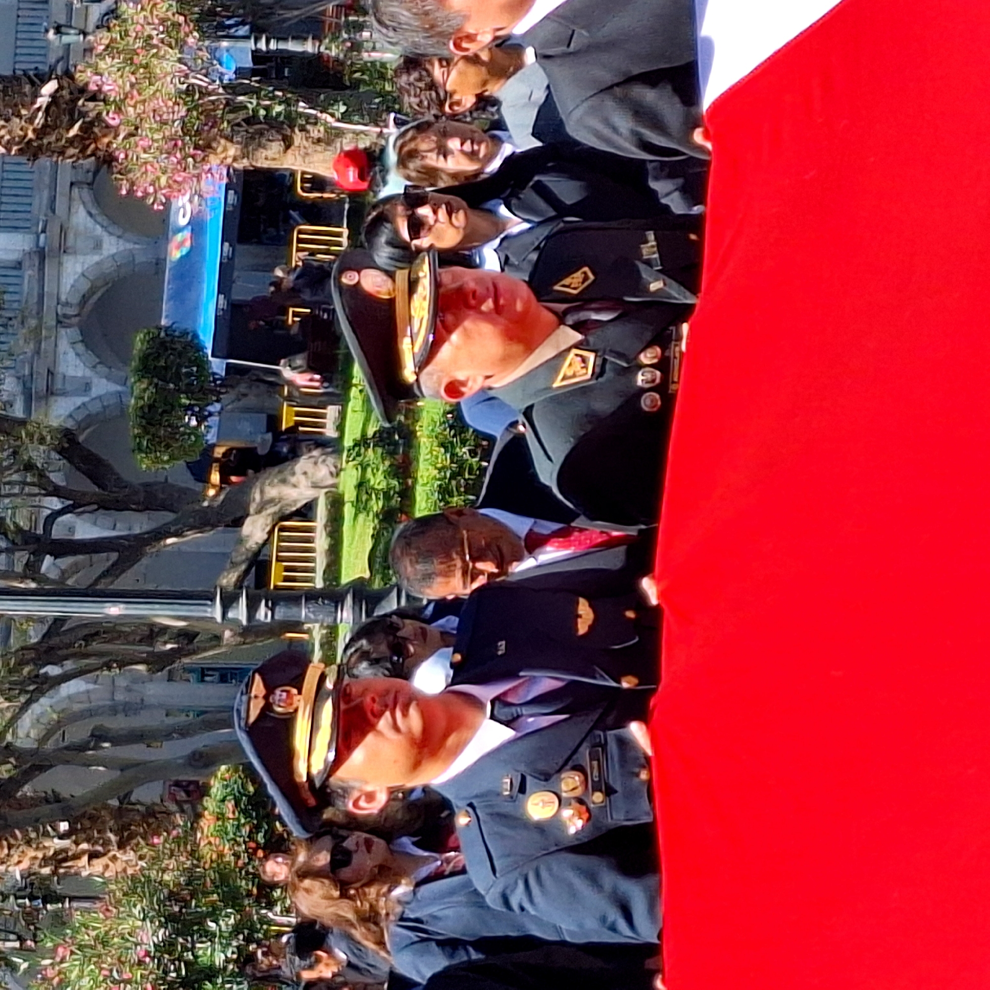 The image shows a group of people, including military officers in uniform, standing in what appears to be a public space with trees and buildings in the background. A large red flag is prominently displayed in the foreground. In the background, the architecture suggests a city setting.