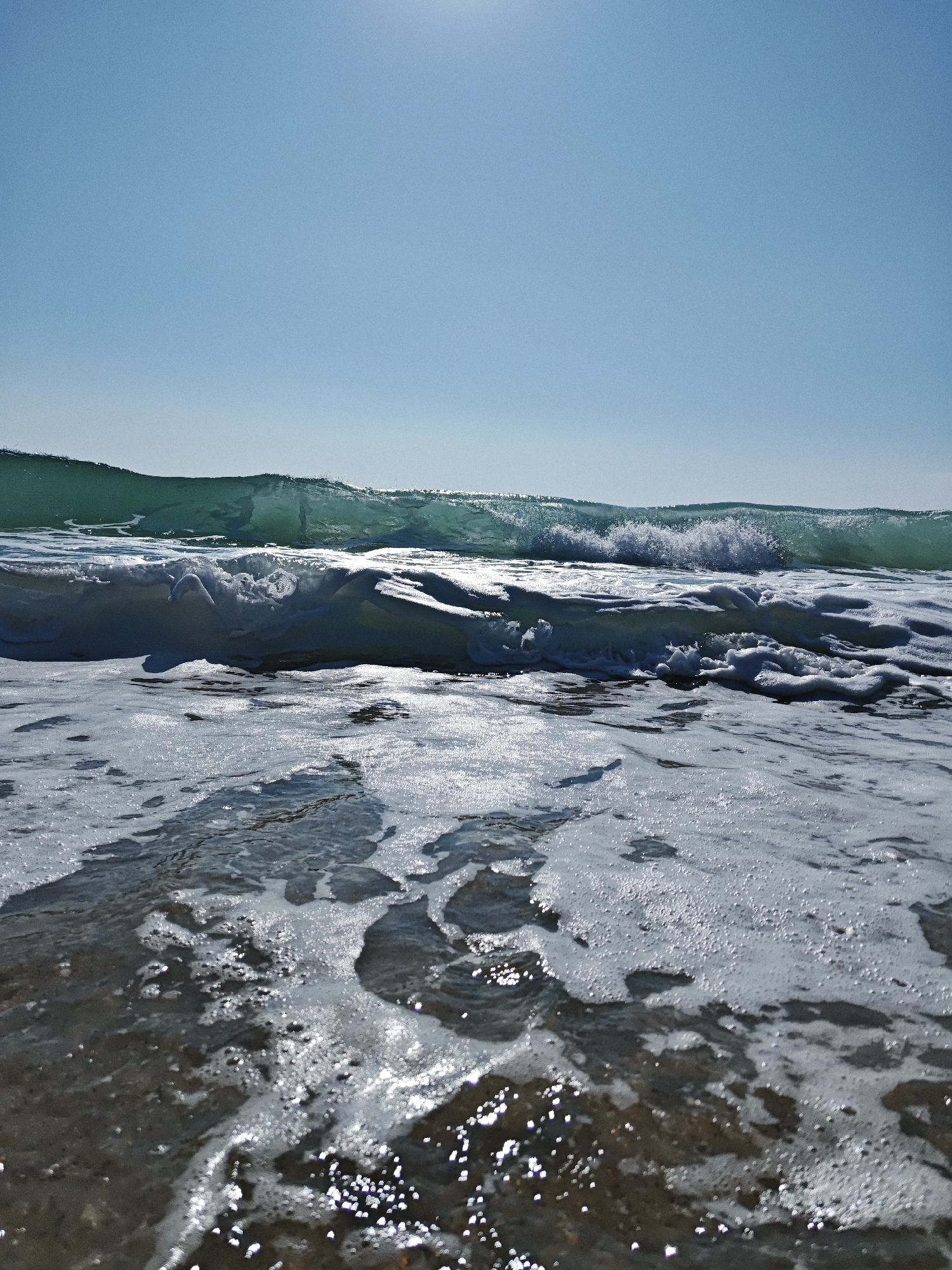A low-angle shot captures an ocean wave breaking under a clear blue sky. The foreground is dominated by frothy white water and shallow, dark sand. The wave itself is a translucent emerald green, with white foam cresting as it curls forward. The sun glints off the water's surface, creating bright highlights. The horizon line is low, emphasizing the vastness of the sky.