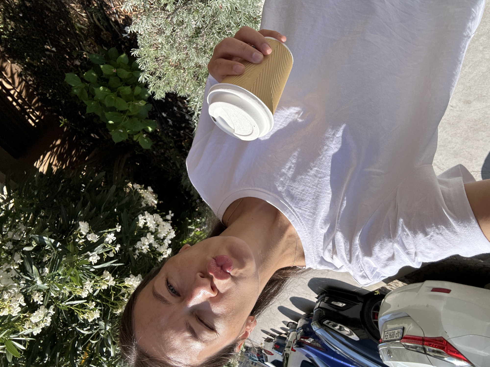 A woman with brown hair is taking a selfie outdoors. She is wearing a white t-shirt and puckering her lips. In her right hand, she holds a disposable coffee cup with a white lid. Behind her, parked cars and green foliage are visible. The image appears to have been taken on a sunny day.