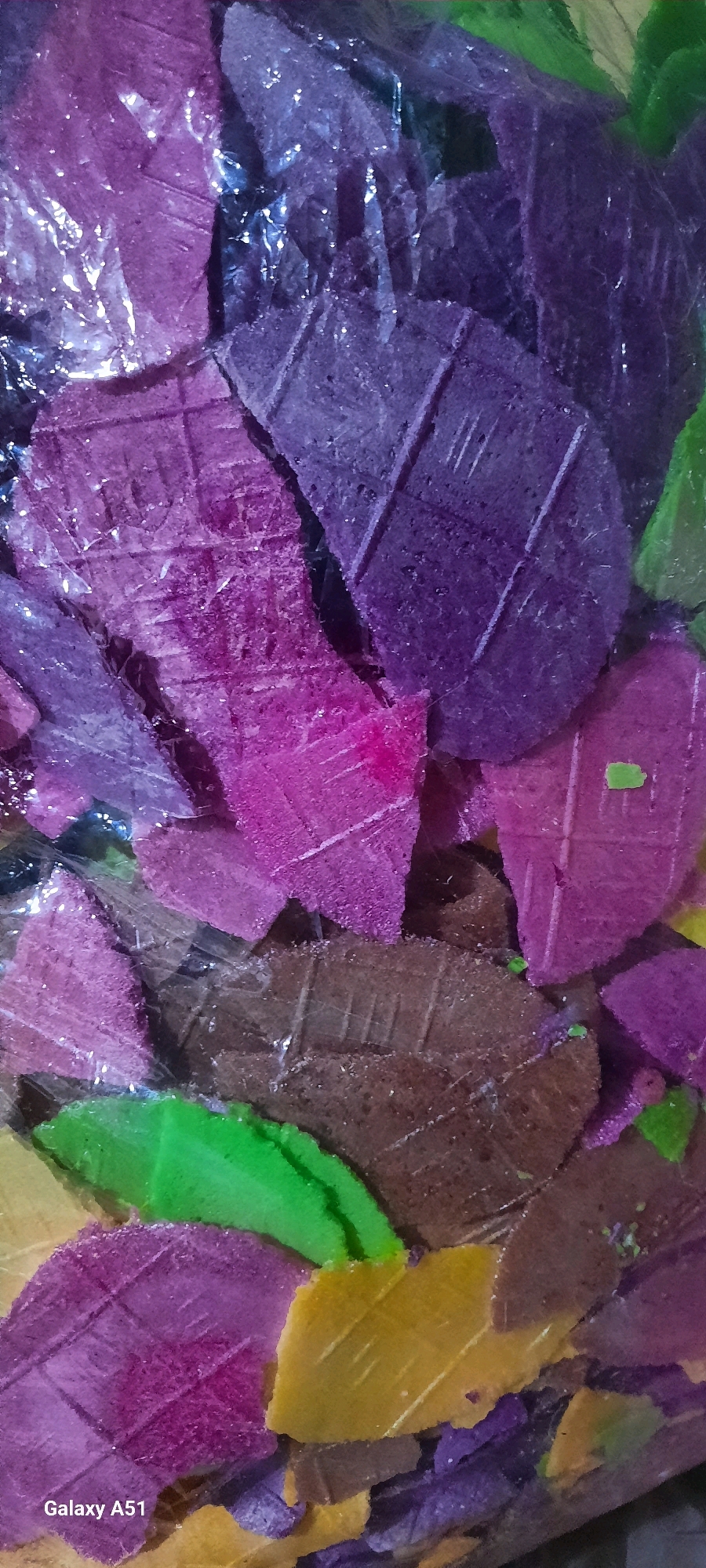 A close-up, vertical, full-frame, high-angle shot shows a pile of brightly colored, flat, irregularly shaped snack pieces. The snacks are predominantly purple, pink, brown, green, and yellow. Many of the pieces have a textured surface with a criss-cross pattern resembling a leaf or a woven material. Some pieces are translucent, suggesting they are wrapped in plastic. The lighting is dim and appears artificial, creating highlights and shadows on the textured surfaces. The bottom left corner of the image displays the text "Galaxy A51", indicating the device used to capture the photograph. The image is likely from Jatitengah, Indonesia, as per the prompt.