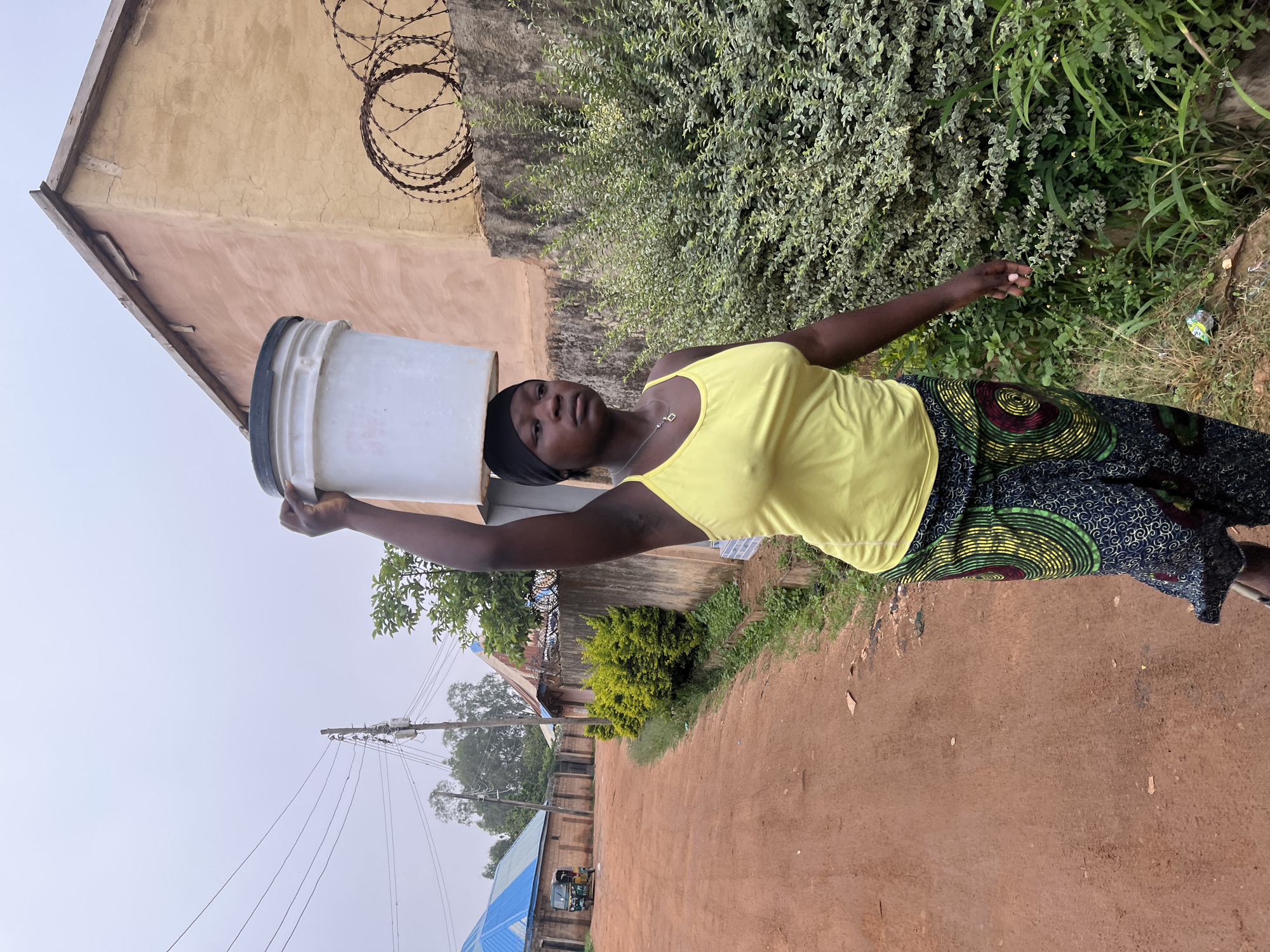 A young woman with dark skin is carrying a large white bucket of water on her head. She is wearing a bright yellow tank top and a patterned skirt with circular designs. She has a black headscarf on. She is standing on a dirt road, with a building to her left and lush green foliage to her right. In the background, there are power lines and more buildings. The sky is overcast, suggesting it is either early morning or late afternoon, or potentially a cloudy day. The overall scene depicts a common activity of water collection in a rural or semi-rural setting. The woman's gaze is directed slightly upwards and to her left, appearing thoughtful or perhaps weary. The environment appears to be warm, typical of Jos, Nigeria.