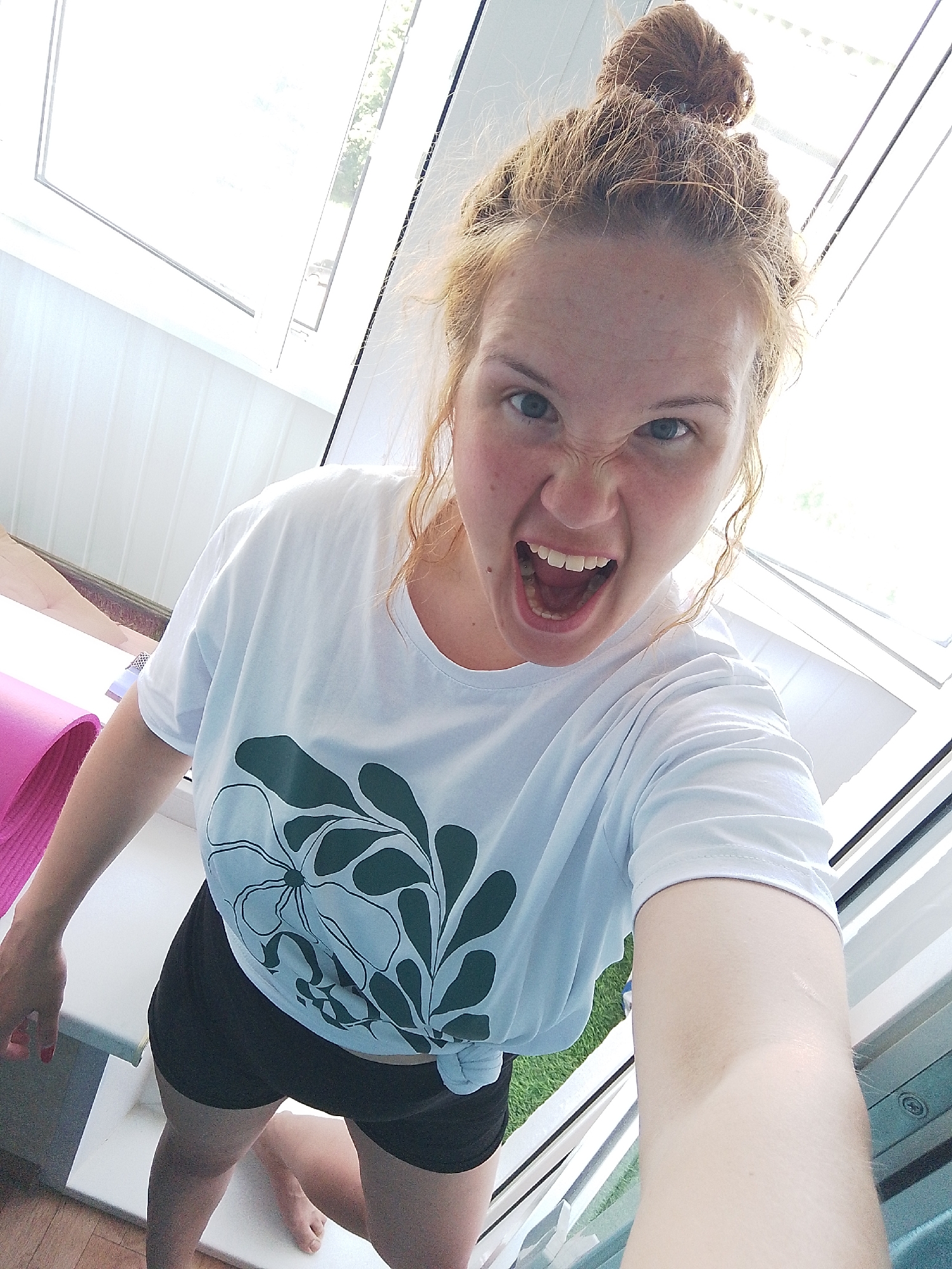 A light-skinned female with red hair styled in a bun is shown in a high-angle selfie. She is wearing a white t-shirt with a dark green floral design and black shorts. The woman is standing with bare feet on a light-colored surface, possibly tile. Behind her, there is a pink yoga mat on a wooden floor. The background includes a window frame and a pane of glass, suggesting the location is indoors, possibly a sunroom or enclosed porch. The lighting is bright, possibly due to sunlight streaming through the windows. The woman is making an aggressive facial expression with her mouth open and teeth bared. The location is not explicitly stated, and therefore it is not known.