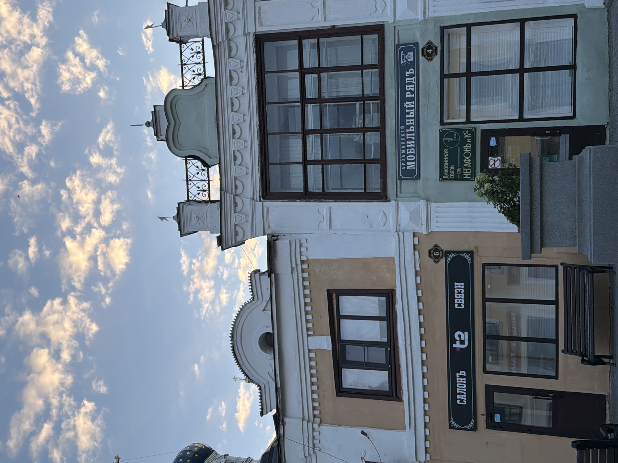 The image captures a street view in Arzamas, Russia, featuring two adjacent buildings under a sky with scattered clouds. The building on the left is painted a pale tan and white, with a prominent dark-framed window above a storefront. The storefront displays a sign with Cyrillic text that translates to "Salon T2 Communication," and a door with glass panes is visible to the left of a large display window. A dark wooden bench is positioned in front of the storefront.
The building on the right is painted a pale green and white. It has a large, dark-framed multi-paned window. Below this window, there are two signs. The upper sign, in Cyrillic, reads "Arzamas Mobile Row" and features a telephone icon. The lower sign displays "Wild Communication - MegaFon and Co" along with a logo. A dark door with glass panes is situated to the left of the display window. A small green bush grows in front of the right building, and a stone planter is present. Another dark wooden bench is visible in the foreground. Architectural details such as decorative cornices and a wrought-iron fence are present on the upper facades of both buildings. The image appears to have been taken during the daytime, possibly late afternoon or early evening, given the lighting conditions.