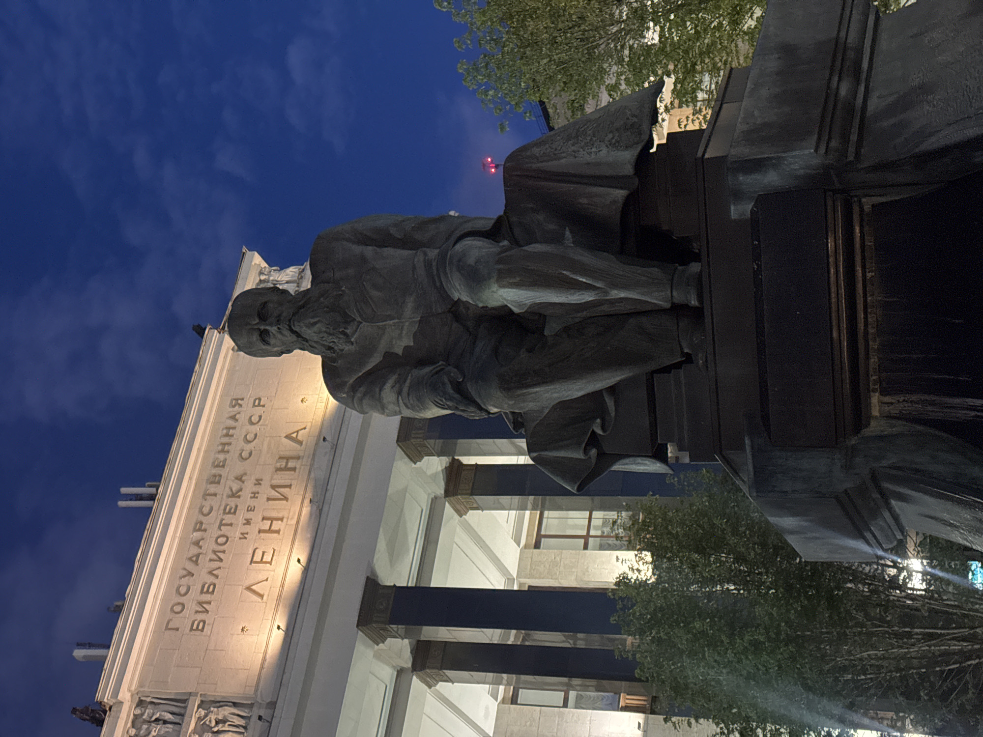 This is a low-angle, nighttime photograph of a large bronze statue of a man with a prominent beard, seated on a pedestal in front of a grand, brightly lit neoclassical building. The building, identified by the prominent Cyrillic inscription "ГОСУДАРСТВЕННАЯ БИБЛИОТЕКА СССР ИМЕНИ ЛЕНИНА" (Lenin State Library of the USSR), is illuminated by spotlights from below, highlighting its ornate facade and columns. The sky above is a deep twilight blue with scattered clouds. The statue itself is detailed, showing the man in a suit with his legs crossed, draped with a cloak. The overall atmosphere is solemn and imposing, likely captured in Moscow, Russia, given the inscription on the building. The statue is a prominent feature in the foreground, with the illuminated library serving as a dramatic backdrop. Trees are visible on the right side of the frame, with their leaves illuminated by the building's lights. The composition emphasizes the grandeur of the architecture and the monumentality of the statue.