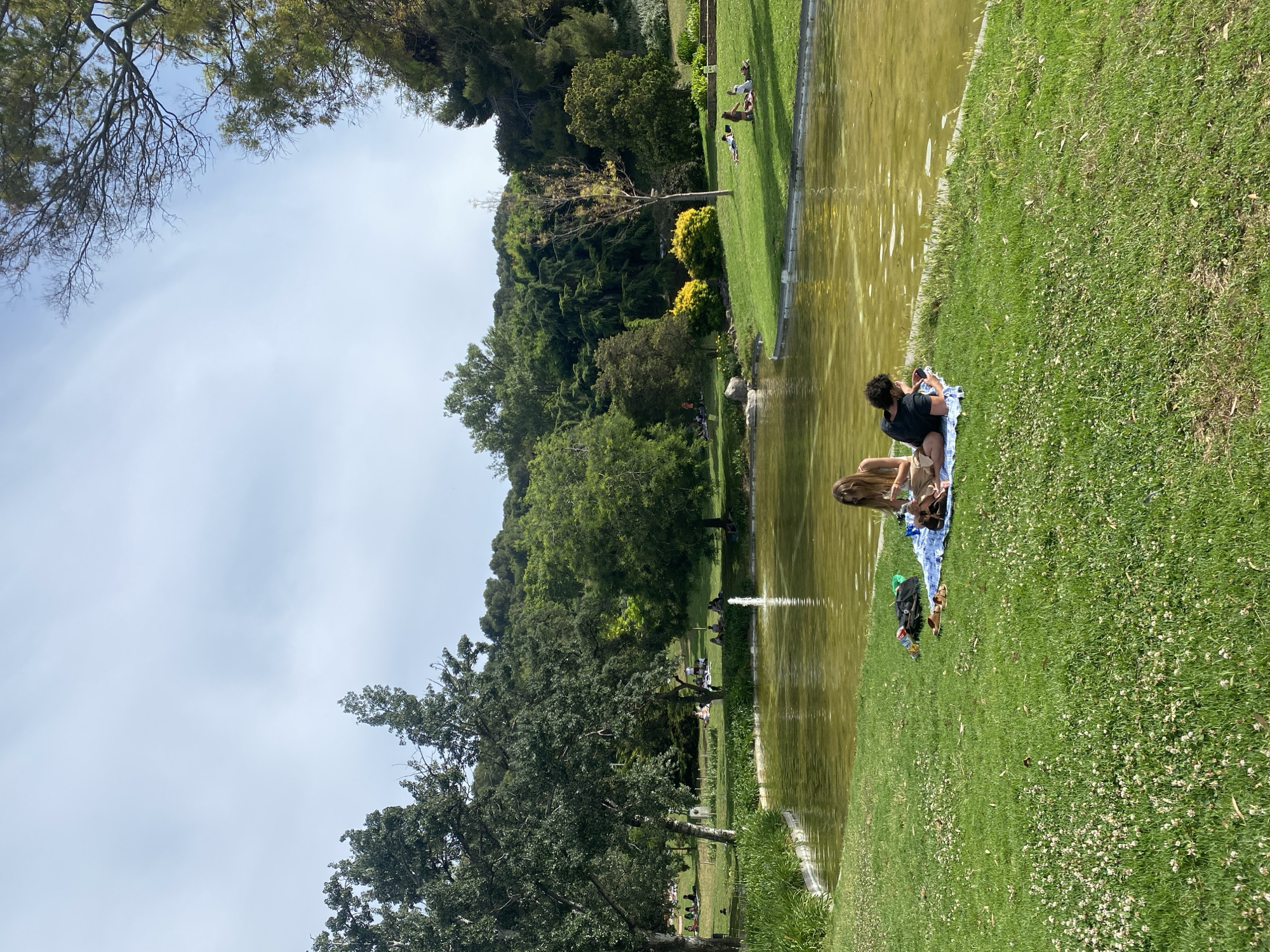 This image captures a wide-angle view of a public park, identified as **Parc de l'Oreneta** in the Sarrià-Sant Gervasi district of Barcelona, Spain, on a brightly lit day.
In the foreground, a verdant grassy area, interspersed with numerous small white clover blossoms, slopes gently towards a pond. Centrally positioned on this grass, two individuals are observed relaxing on a blue and white striped blanket. The individual on the left, with long brown hair, is lying on their stomach, propped on their elbows, wearing a light-colored top and light-colored trousers. A dark backpack and light brown sandals are situated beside them. The individual on the right, a male with dark hair, is wearing a black t-shirt and dark shorts, lying on his side and appearing to engage with a device in his hands.
The midground features a calm, olive-green pond with subtle reflections on its surface. A single fountain on the far side of the pond emits a vertical stream of water. The pond's edges are lined with either natural earth or subtle concrete/stone barriers, and a section on the left bank is dense with tall green reeds or grasses. Beyond the pond, extensive grassy slopes rise, populated by multiple small groups and individuals scattered across the landscape, many seated or lying on the grass or blankets. Various trees with both light and dark bark, and dense green foliage, are distributed throughout this area, providing shade.
The background is dominated by a substantial, densely forested hill, characterized by a mix of evergreen and deciduous trees, indicating the park's integration with a natural, elevated landscape. The sky above is a pale blue, with wisps of scattered white clouds. The overall lighting suggests natural sunlight, creating relatively soft shadows.