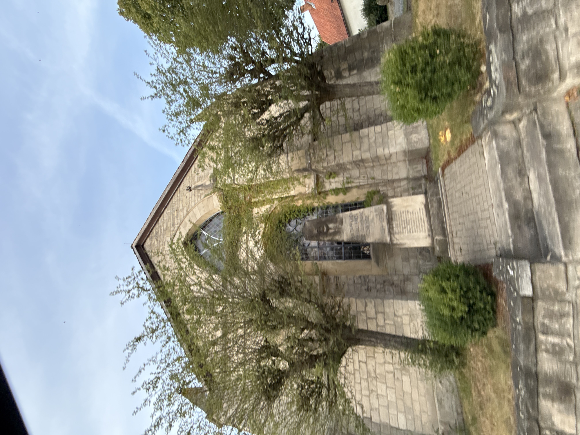 The image shows the exterior of a stone church in Bad Sachsa, Germany, likely St. Nicholas' Church given its architectural style and the presence of a war memorial. The church features a large arched window and is partially covered in ivy. In front of the church, there is a war memorial with inscriptions, a paved walkway, and manicured bushes. The sky above is blue with some wispy clouds, and trees with green foliage frame the scene. The overall impression is that of a serene, historical religious site.