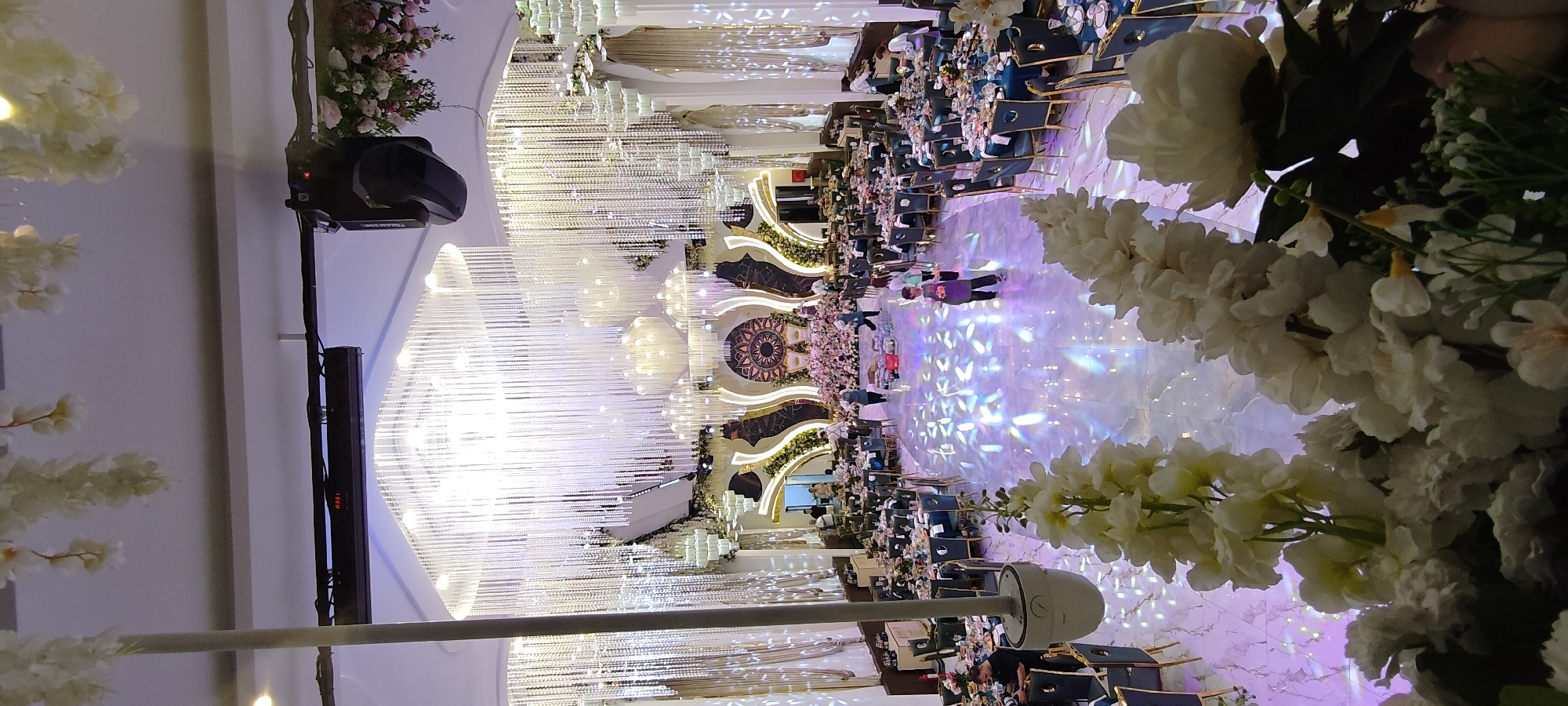 The image depicts a spacious indoor venue, likely a banquet hall or event space, in Khujand, Tajikistan. The hall is elaborately decorated, featuring large, multi-tiered chandeliers with numerous crystal-like strands that illuminate the space. The walls are adorned with ornate gold and dark patterned designs, and large, flowing drapes hang from the upper portions of the walls. A central stage area is decorated with an abundance of pink flowers and features two ornate chairs, suggesting a ceremonial or celebratory event.
The floor is a polished white marble, reflecting the light from the chandeliers and projecting a pattern of purple and white light across its surface. Round tables, covered with dark blue tablecloths and set with tableware, are arranged throughout the hall, indicating a dining or seating arrangement for guests. Some tables are laden with food and drinks.
Several individuals are visible in the scene. In the foreground, on the right side, large white floral arrangements are prominently displayed, partially obscuring the view. Towards the center and middle ground, people are seen walking around, some appearing to be staff members preparing or serving. One individual in the middle distance is wearing a purple apron and carrying something, suggesting they are part of the event staff. Another person is visible in the left middle ground, appearing to be a photographer with a camera. A group of people are also seen near the entrance area on the left. The overall atmosphere appears to be one of preparation or the early stages of an event. The lighting is bright and theatrical.