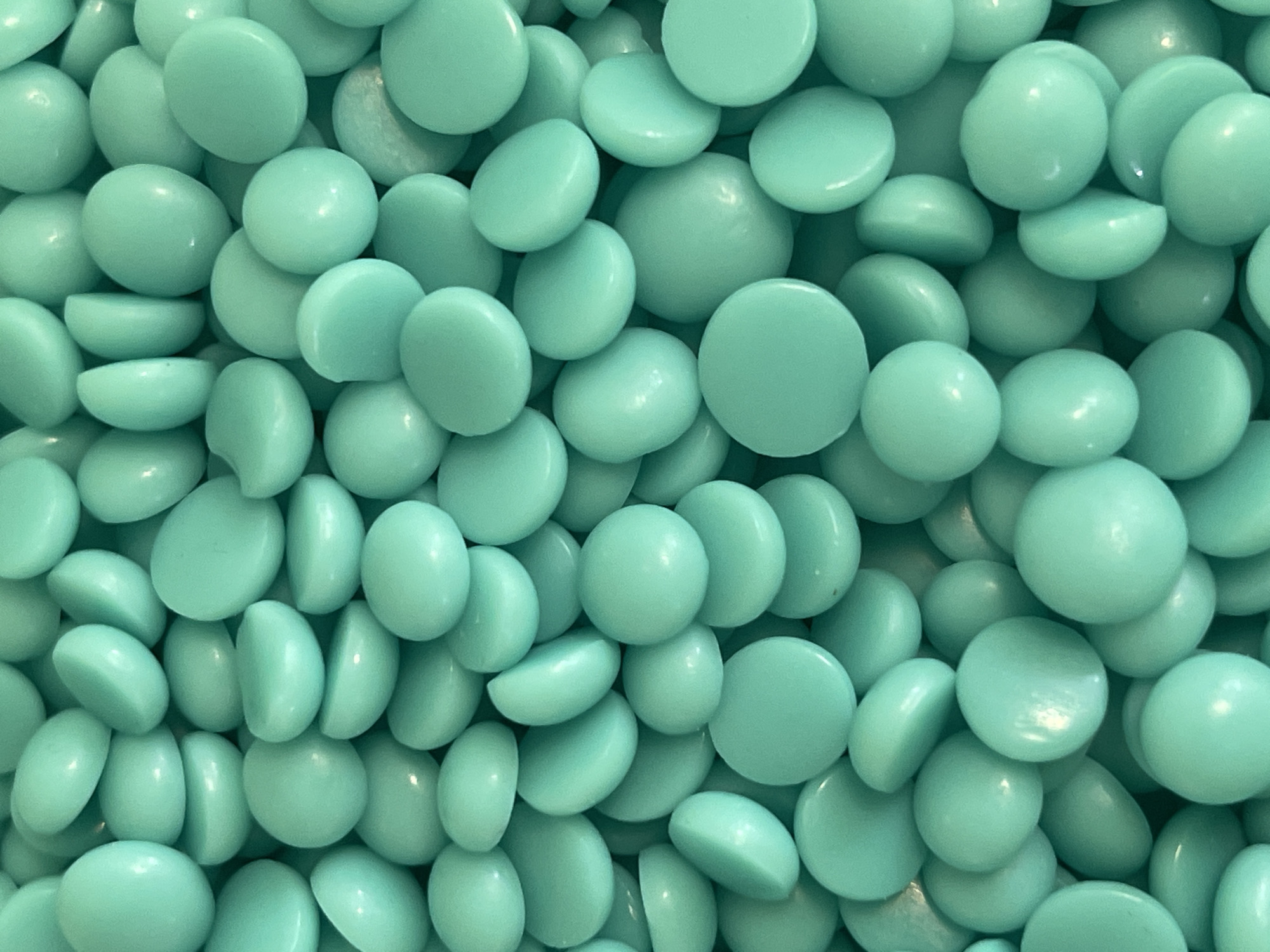 The image displays a tightly packed, overhead view of numerous small, disc-shaped, teal-colored beads. The beads are varied in their orientation and overlap, creating a dense texture. Some beads appear to be whole, while others are cut in half, revealing their smooth, rounded interior. The lighting highlights the glossy surface of the beads, creating soft shadows and reflections. The scene is uniform, with no discernible background or context beyond the beads themselves. There is no indication of a specific location in Kyiv, Ukraine, as the image is a close-up of a manufactured object.