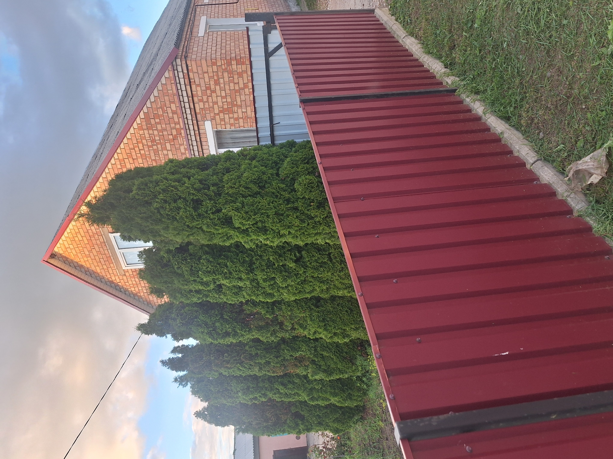 The image depicts a residential property in the Капыльскі сельскі Савет, Belarus, under a cloudy sky with hints of sunlight. In the foreground, a red corrugated metal fence runs horizontally across the frame, with a dark vertical support post visible on the left. Behind the fence, a row of tall, green evergreen trees stands in a line. Beyond the trees, a two-story brick house is visible, featuring a red-tiled roof and white-framed windows. A section of a light-colored metal fence is also present behind the evergreen trees. The ground is covered with grass and some debris, including a crumpled plastic bag. The overall scene suggests a suburban or rural residential setting.