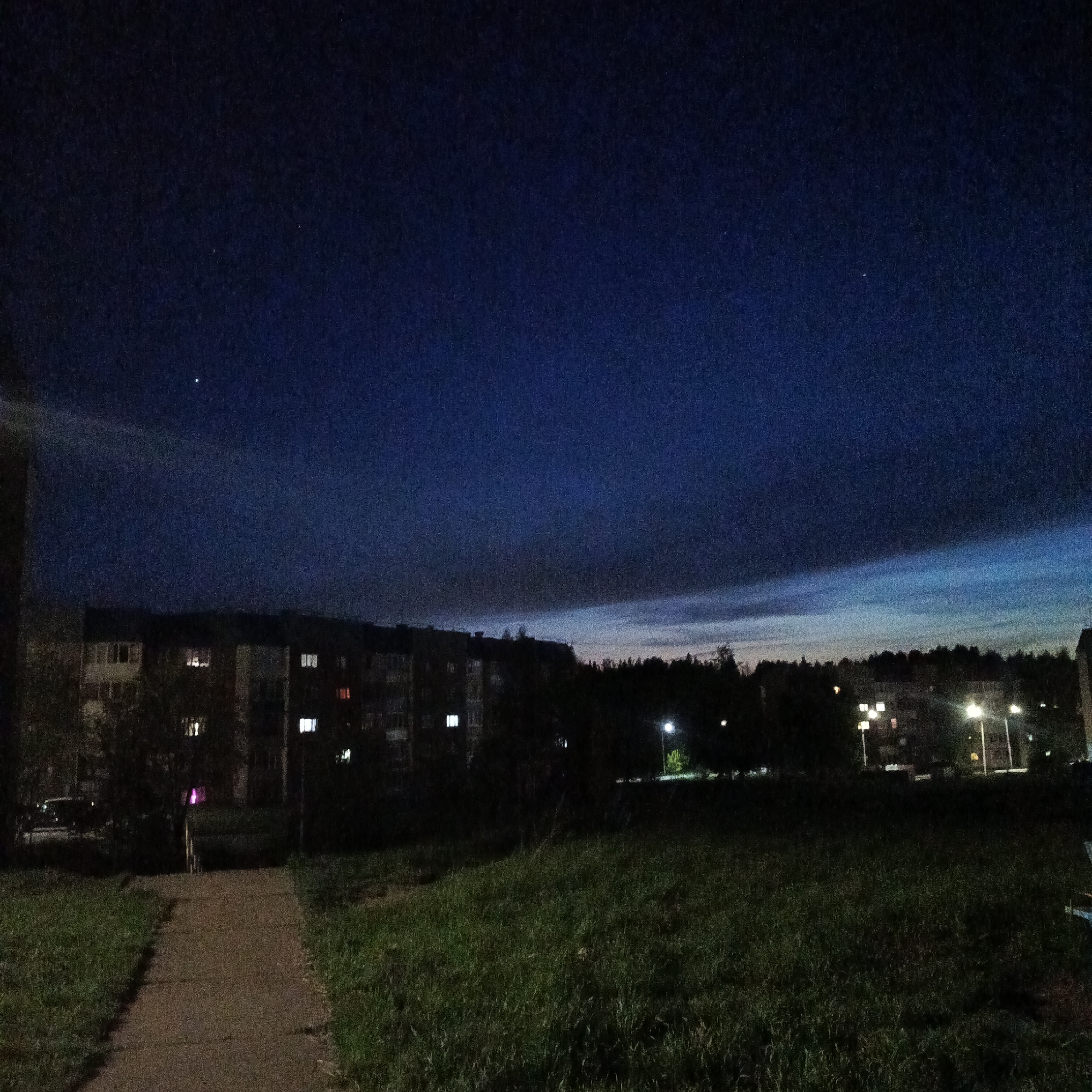 The image depicts an outdoor scene in Markovskiy, Russia, at dusk or night. The sky is a gradient of dark blue to lighter blue towards the horizon. There are a few visible stars. In the midground are apartment buildings with some illuminated windows. Streetlights are present, casting light onto the vegetation. The foreground shows a grassy area and a concrete walkway. No people are visible.