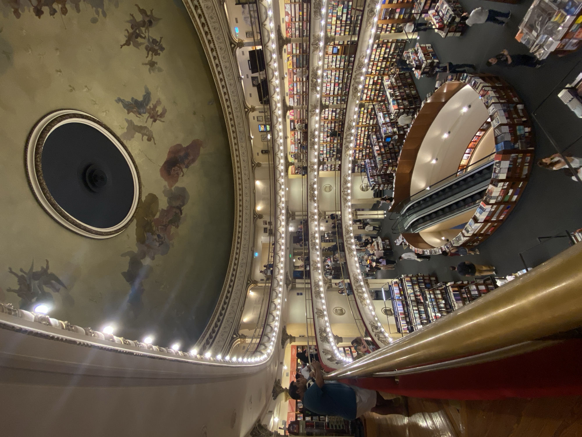 This image depicts the interior of El Ateneo Grand Splendid bookstore, located at Santa Fe Avenue 1860 in the Recoleta neighborhood of Buenos Aires, Argentina. The scene is a wide-angle view from an elevated balcony, looking down into a converted grand theater.
The multi-tiered structure features an ornate, high arched ceiling adorned with a fresco painting depicting numerous ethereal, cherubic figures in flight against a beige-green sky. A large, dark oval opening is centered in the ceiling. Below, five visible levels of curved balconies, originating from the former theater boxes, are illuminated by rows of small, downward-facing lights. Each balcony railing is a dark red, topped with a polished gold handrail, and features intricate white and gold decorative moldings.
The central area of the ground floor, beneath the balconies, is occupied by extensive bookshelves arranged in rows and around various display tables. Two parallel escalators descend into a lower, oval-shaped sunken area also lined with books. Thousands of books are visible on shelves spanning all levels and on freestanding displays.
Approximately 30-40 individuals, primarily adults, are distributed throughout the bookstore. A male subject in a blue shirt and white shorts leans on the golden railing in the immediate foreground, looking left. Other individuals are seen browsing books on various levels, walking through aisles, or standing near the escalator and checkout areas on the ground floor. The overall lighting is bright and warm, accentuating the architectural details and the vast collection of books.