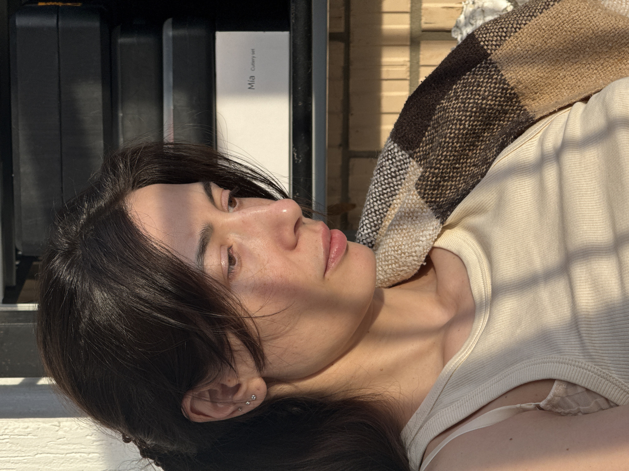 A medium close-up shot depicts a person with dark brown hair and fair skin, lying down in what appears to be an outdoor setting. The person is oriented on their right side. A section of a plaid blanket in shades of brown and beige is visible above the person. The person is wearing a light-colored tank top and earrings in the left ear. Shadows fall across the person's face and upper body.
Behind the person is a set of black, rectangular objects next to a white box labeled "Mia Cutlery Set". Beyond these objects, a brick wall can be seen. The city is unknown.