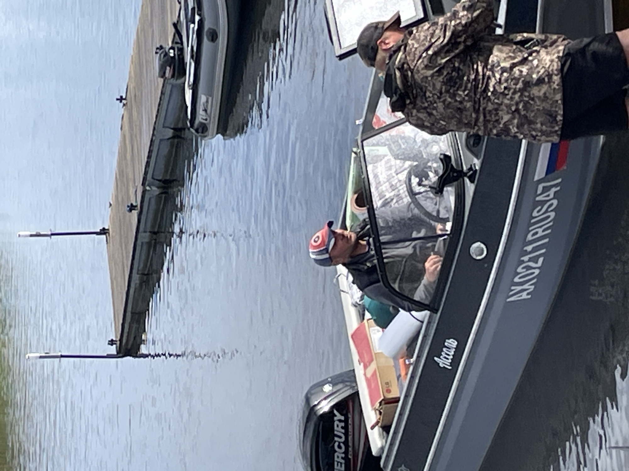 Two individuals are in a gray motorboat on a body of water, likely a river, near a wooden dock. The boat has "Mercury" branding on its outboard motor and the registration number "AX0211RUS47" on its hull. The boat is stocked with various items including boxes, a white rolled object, and green bags. The person in the driver's seat is wearing a cap and a dark vest. Another person, wearing a camouflage jacket and a black cap, is standing in the boat. In the background, an inflatable boat with an outboard motor is tied to the dock. The water is calm with reflections of the sky and dock visible. The scene suggests a leisurely activity or fishing trip in Berozovo, Russia, given the registration number.