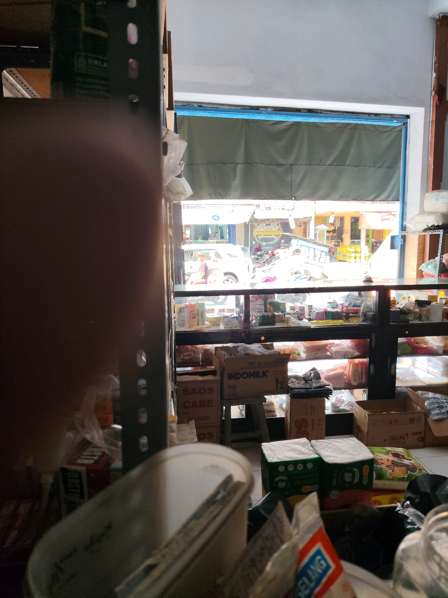 The image depicts the interior of a small shop or minimarket, likely in Dampit, Indonesia, as suggested by the context. The foreground is dominated by the left side of a large, out-of-focus object, possibly a shelf or a refrigerator, with some blurry plastic bags and containers in front of it. To the right, a display cabinet is visible, filled with various packaged goods such as bottles of liquid, boxes, and jars. These items appear to be toiletries, food products, and possibly some over-the-counter medications.
Through a large opening or window to the back of the shop, an outdoor scene is visible. The outdoor environment suggests a street with buildings and vehicles. There are several motorcycles parked, a car, and what appears to be a food cart or a small stall. People are present in the outdoor scene, including individuals riding motorcycles and standing. The overall lighting suggests it is daytime, with sunlight filtering into the shop from the outside. The shop itself seems somewhat cluttered, with items on shelves and on the floor, indicating a busy retail environment.