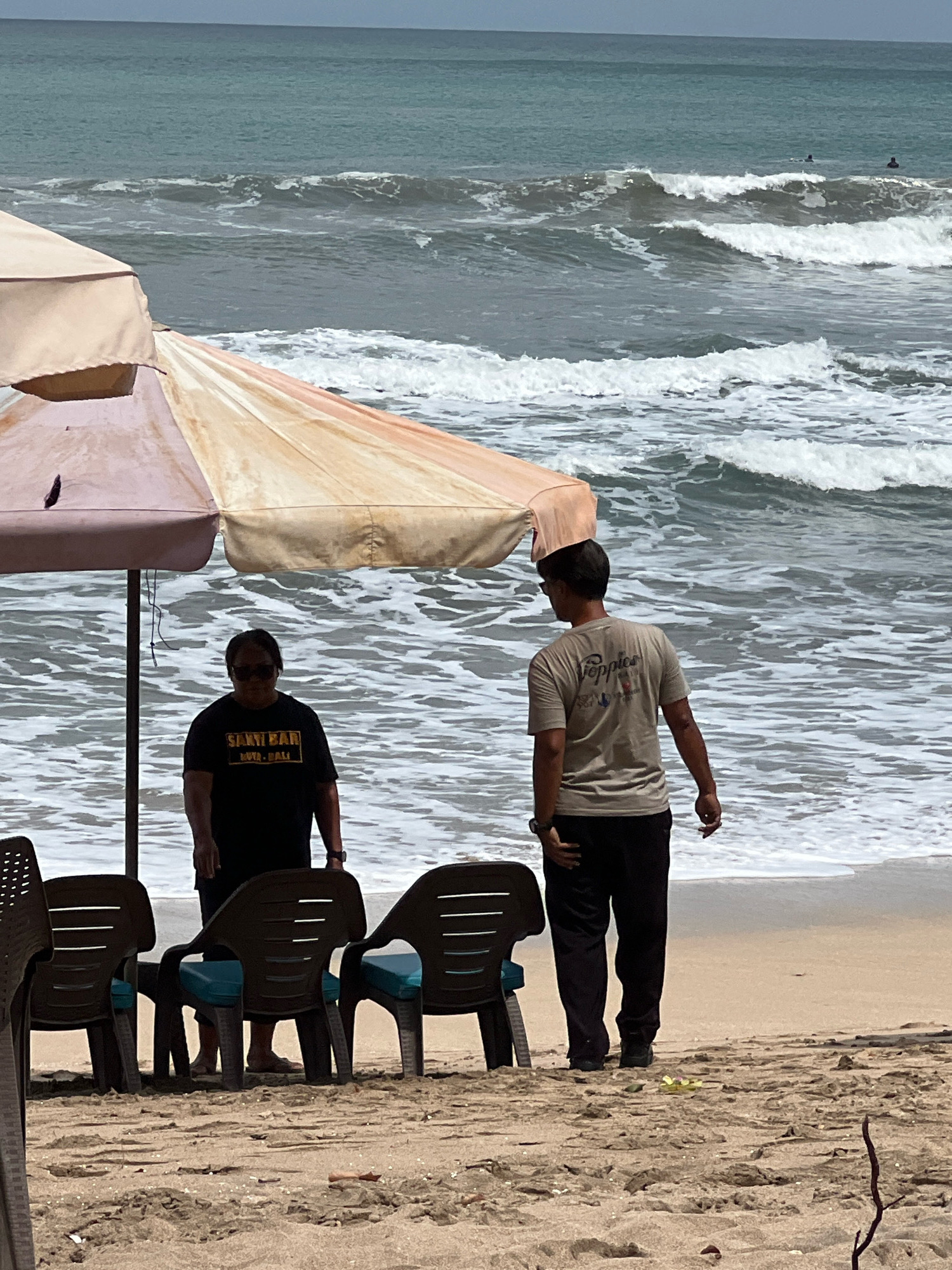 Two individuals are on a sandy beach in Kuta, Indonesia, near the ocean. A large, multi-colored beach umbrella shades a portion of the sand. In the foreground, three dark plastic beach chairs with blue cushions are arranged. To the left of the chairs, a person wearing sunglasses and a black t-shirt with "SANTRI BAR" and "LUX - BALI" printed on it stands with their back to the camera, facing the ocean. To the right, a person wearing a tan t-shirt with a "Poppies" logo and dark pants stands with their back to the camera, also facing the ocean. The ocean in the background displays moderate waves and several distant surfers. The sky is overcast.