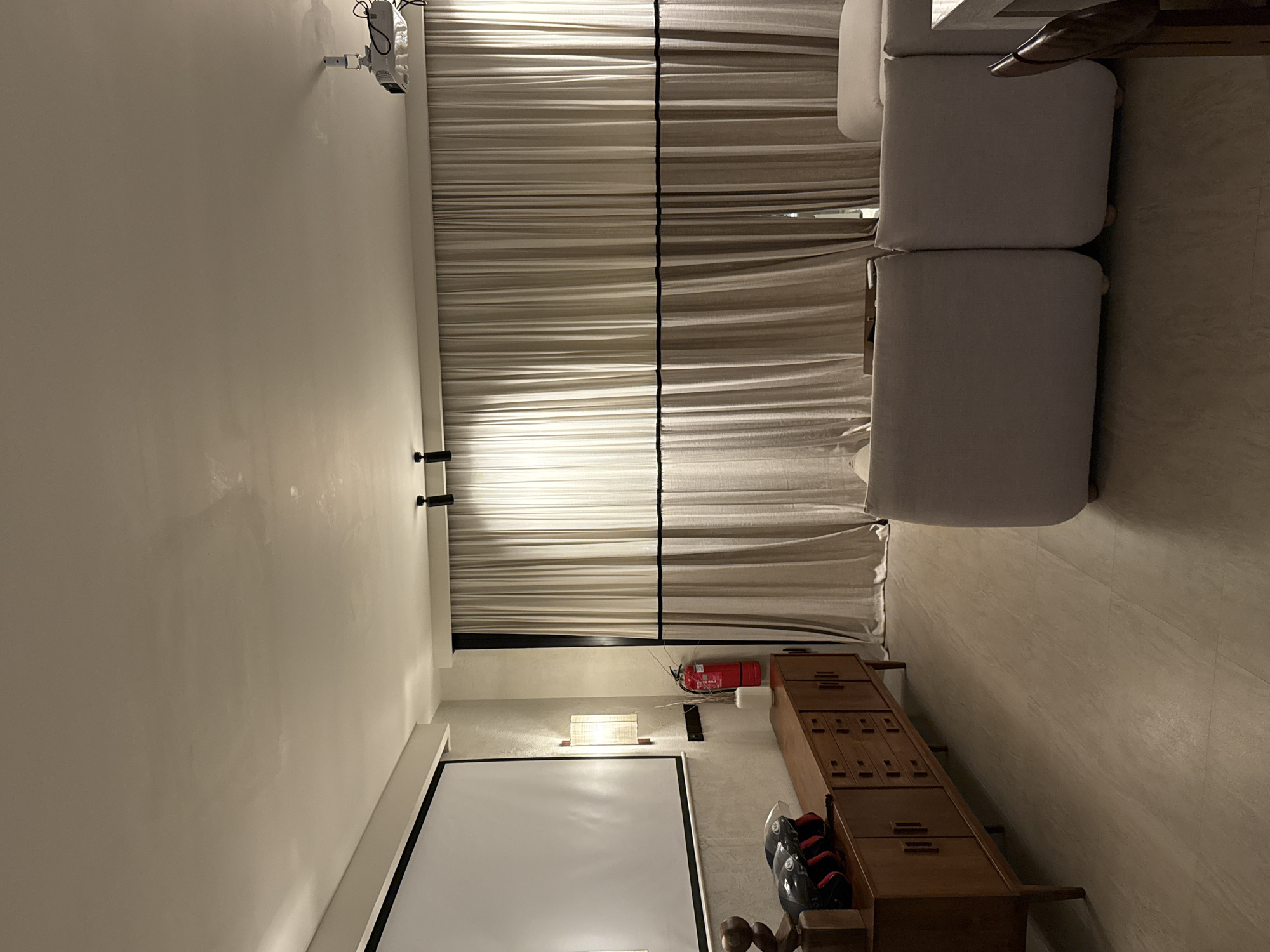 The image depicts an interior space, possibly in a residential or entertainment setting. A white projector screen is visible on the left side of the frame. Above the screen is a wall-mounted light fixture, a fire extinguisher, and a long, wooden cabinet with drawers. A row of black and red sports helmets sits on top of the cabinet. The background features draped curtains, and a projector is mounted on the ceiling. A section of a light-colored sofa is visible to the right of the curtains.
The location is presumed to be in the city of Kuta Selatan, Indonesia, though this is based on external information. No further identifying information is available from the image.