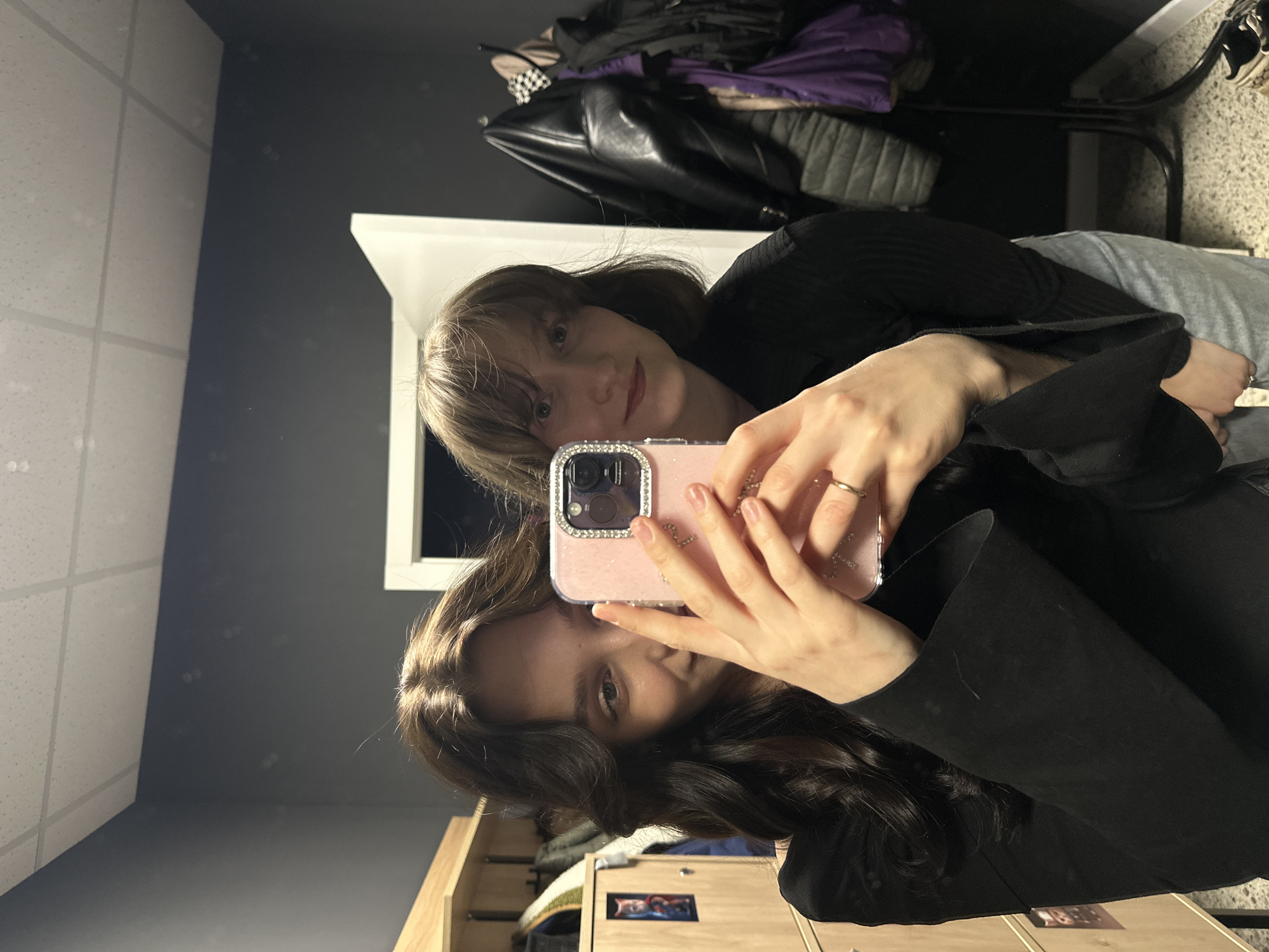 Two women are taking a selfie in a room. The woman in the foreground has dark hair and is wearing a black shirt or jacket. She is holding up a pink phone with a jeweled case. The woman in the background has light brown hair with bangs and is also wearing black. They are standing in front of a dark gray wall with white ceiling tiles. In the background, there is also a rack of clothing with a black leather jacket and purple fabric. The location is Unknown, Unknown.