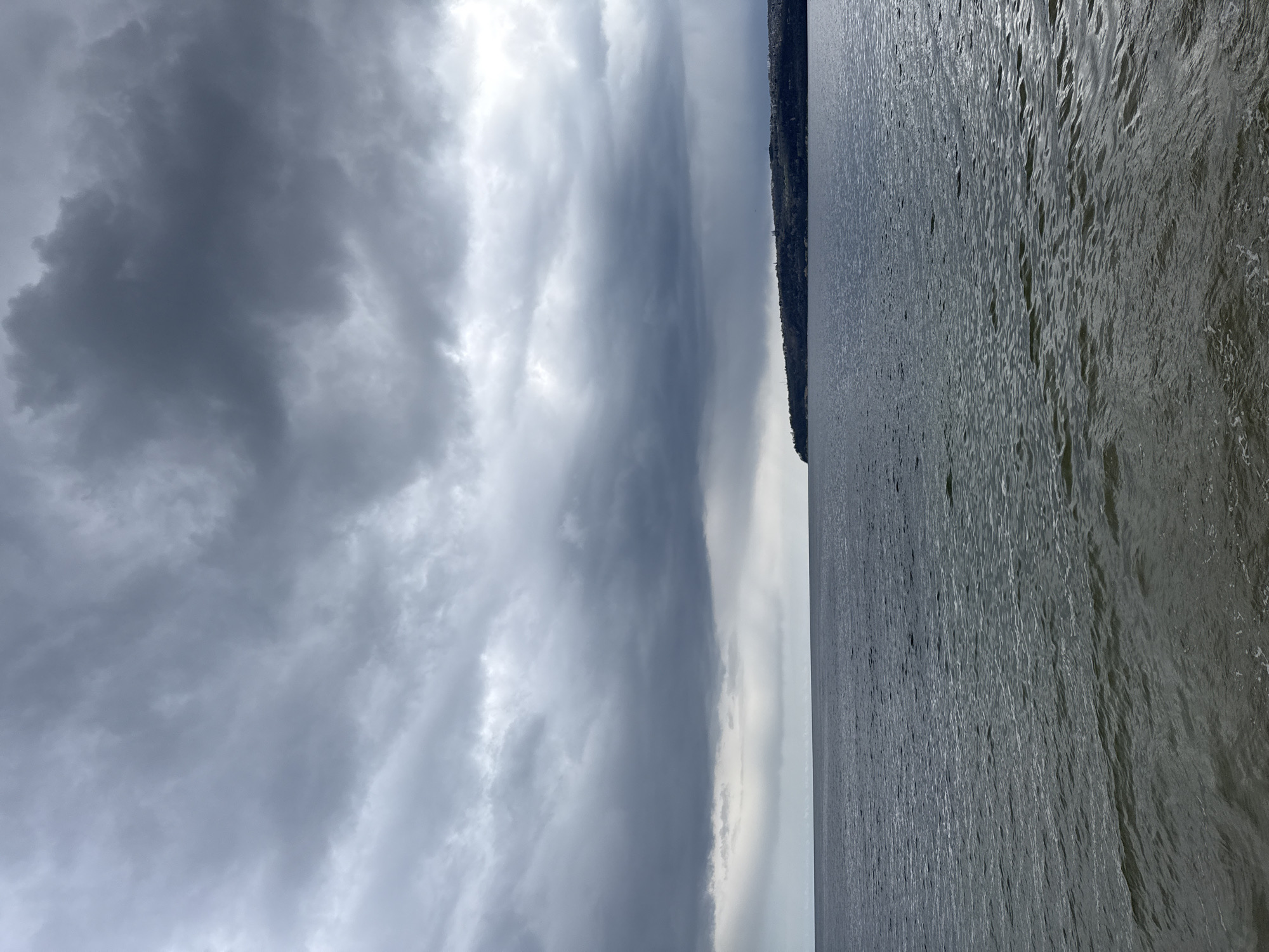 The image presents a seascape under a heavily clouded sky. To the right, the water, likely a part of the Black Sea near Varna, Bulgaria, shows a rippled surface. A strip of land with dense vegetation is visible along the horizon line, possibly indicating the coastline or a nearby island. The dominant feature is the sky, filled with layers of dark, ominous clouds, suggesting an overcast or stormy weather condition. There are no visible people or signs of human activity apart from the shoreline.