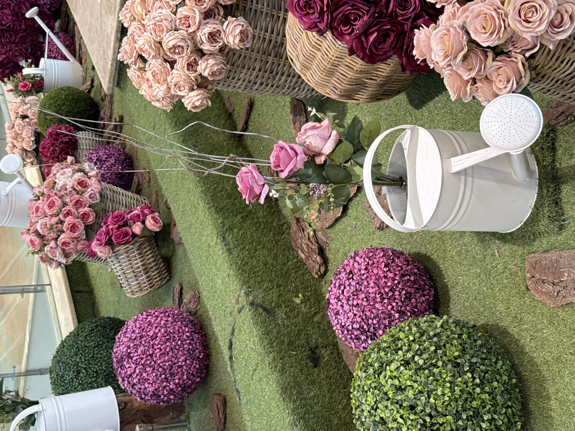 This image captures a vibrant and meticulously arranged artificial garden or floral display, viewed from an overhead perspective looking down onto the scene. The ground is covered with a lush artificial green grass, creating a consistent and inviting backdrop.
Dominating the display are numerous arrangements of artificial roses in various stages of bloom. These flowers showcase a rich palette of colors, including soft dusty pinks, delicate creams, and deep, luxurious magenta and burgundy hues. Many of these bouquets are held within rustic woven wicker baskets of different sizes, adding a natural, handcrafted element to the setup.
Several decorative watering cans are strategically placed throughout the scene. A prominent white or light gray metal watering can with a distinct sprinkler head is positioned towards the right side of the frame, appearing to lie on its side. Other smaller watering cans are partially visible elsewhere, all serving as charming props.
Interspersed among the floral arrangements and grass are spherical, topiary-like decorations. These come in two primary colors: a vibrant green, mimicking manicured shrubs, and a striking fuchsia or purple, suggesting flowering bushes. Pieces of natural wood bark and slender, light-colored decorative branches are also scattered across the artificial grass, enhancing the garden aesthetic.
The scene suggests an indoor setting, likely a retail display in a flower shop, a home decor store, or a decorative setup for an event. The bright and even lighting further indicates an indoor environment. There are no people visible, and no discernible text. The overall impression is one of beauty, springtime, and curated decorative charm.