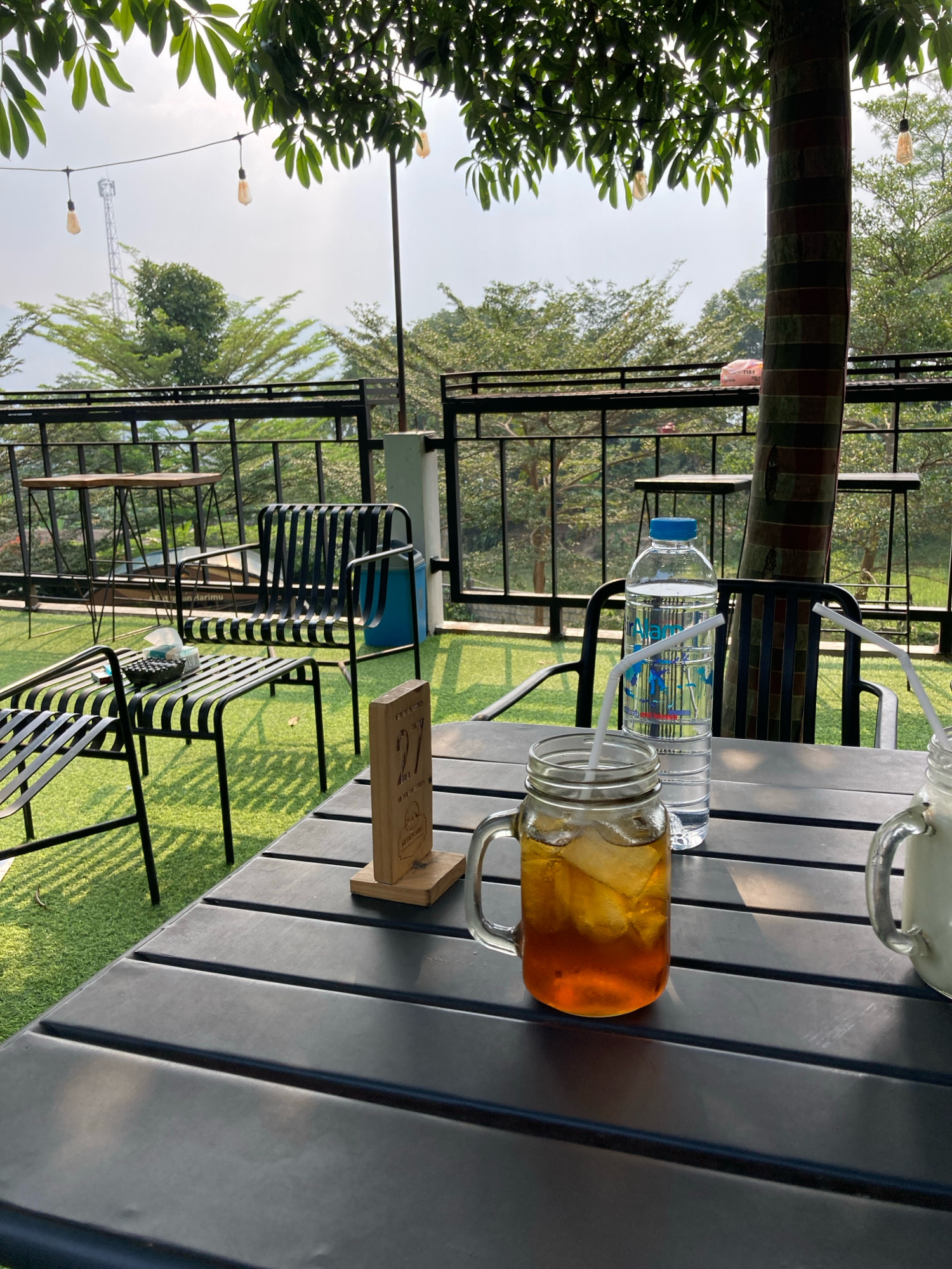 This image captures a close-up of a table with drinks at an outdoor cafe in Sukamakmur, Indonesia. The table is dark and made of horizontal slats, with artificial turf covering the ground. On the table, a glass mason jar filled with iced tea and a straw is prominently featured in the foreground. To its right, another glass, partially visible, appears to contain a white beverage. Behind the iced tea, a bottle of water with a blue cap and a straw is standing upright. A small wooden sign with the number "27" is placed on the table near the iced tea. In the background, black metal chairs and tables are visible, as well as a black metal railing and a lush green landscape with trees. String lights with Edison bulbs are strung across the top of the frame, and a cell tower can be seen in the distance against a hazy sky. The overall setting suggests a casual dining or relaxation spot.