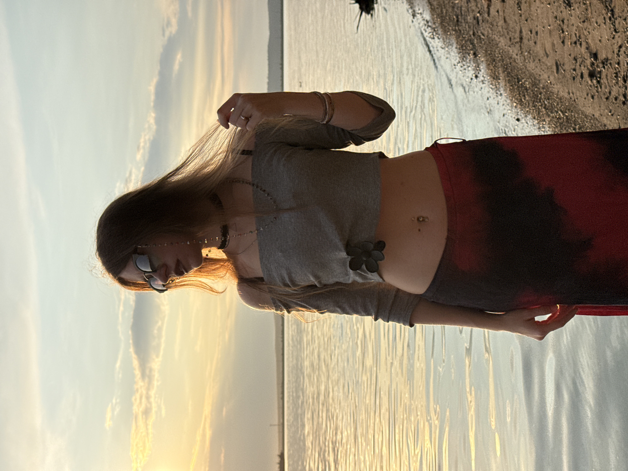 A young woman stands at the edge of a body of water, facing right, at sunset. The sky is a pale yellow and blue, with streaks of clouds. The water is calm and reflects the sunset. The woman has long, brown hair and is wearing sunglasses, a grey off-the-shoulder crop top, and a long, dark red and black skirt. She has a belly button piercing and is wearing jewelry, including a necklace and bracelets. Her right hand is raised, and she is playing with her hair. She is standing on a sandy shore with pebbles. The scene appears to be a natural setting, possibly a beach or a lake.