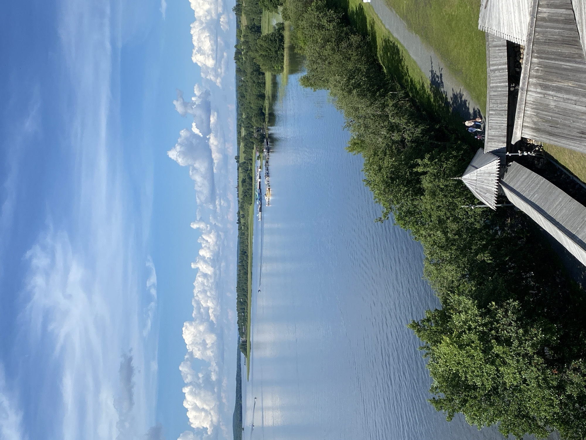 The image captures a scenic view of a wide, tranquil lake on a bright, sunny day in Boyarshchina, Russia. The sky is a vivid blue, dotted with fluffy white clouds. The lake, reflecting the sky, stretches towards a distant tree-lined shore. Several small boats and a dock with moored vessels are visible on the water. In the foreground, a pathway winds through a grassy area bordered by lush green trees. Wooden structures, possibly part of an architectural ensemble, with characteristic roofs and wooden walkways, are prominent in the lower right portion of the image. Two individuals, a child and two adults, are walking along the pathway. The overall impression is one of a peaceful, natural setting with elements of human activity and historical architecture.