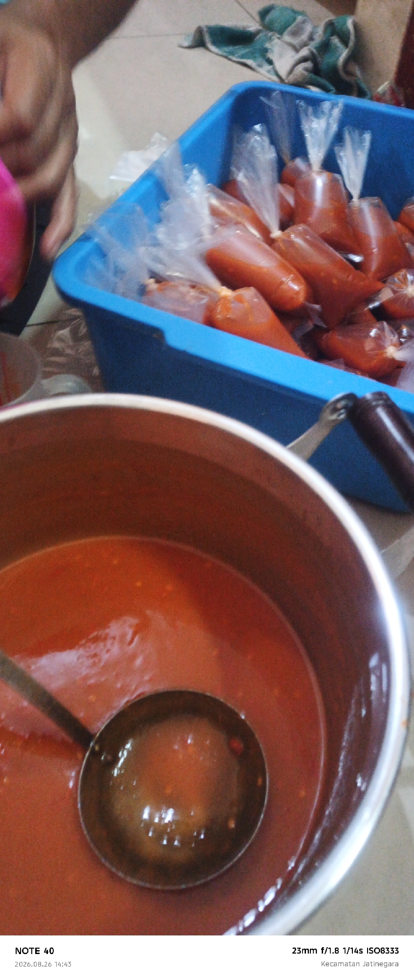 In the foreground, a large metal pot contains a red sauce or soup. A ladle is partially submerged in the liquid. In the background, a blue plastic container is filled with small plastic bags, each containing a portion of the same red sauce. A hand is visible on the left side of the frame, holding a pink object. The scene appears to be indoors, with a tiled floor visible. The bottom of the image includes text indicating "NOTE 40", a date "2026.08.26", a time "14:43", camera settings "23mm f/1.8 1/14s ISO8333", and a location "Kecamatan Jatinegara". This suggests the image was taken in the Jatinegara district of East Jakarta, Indonesia.