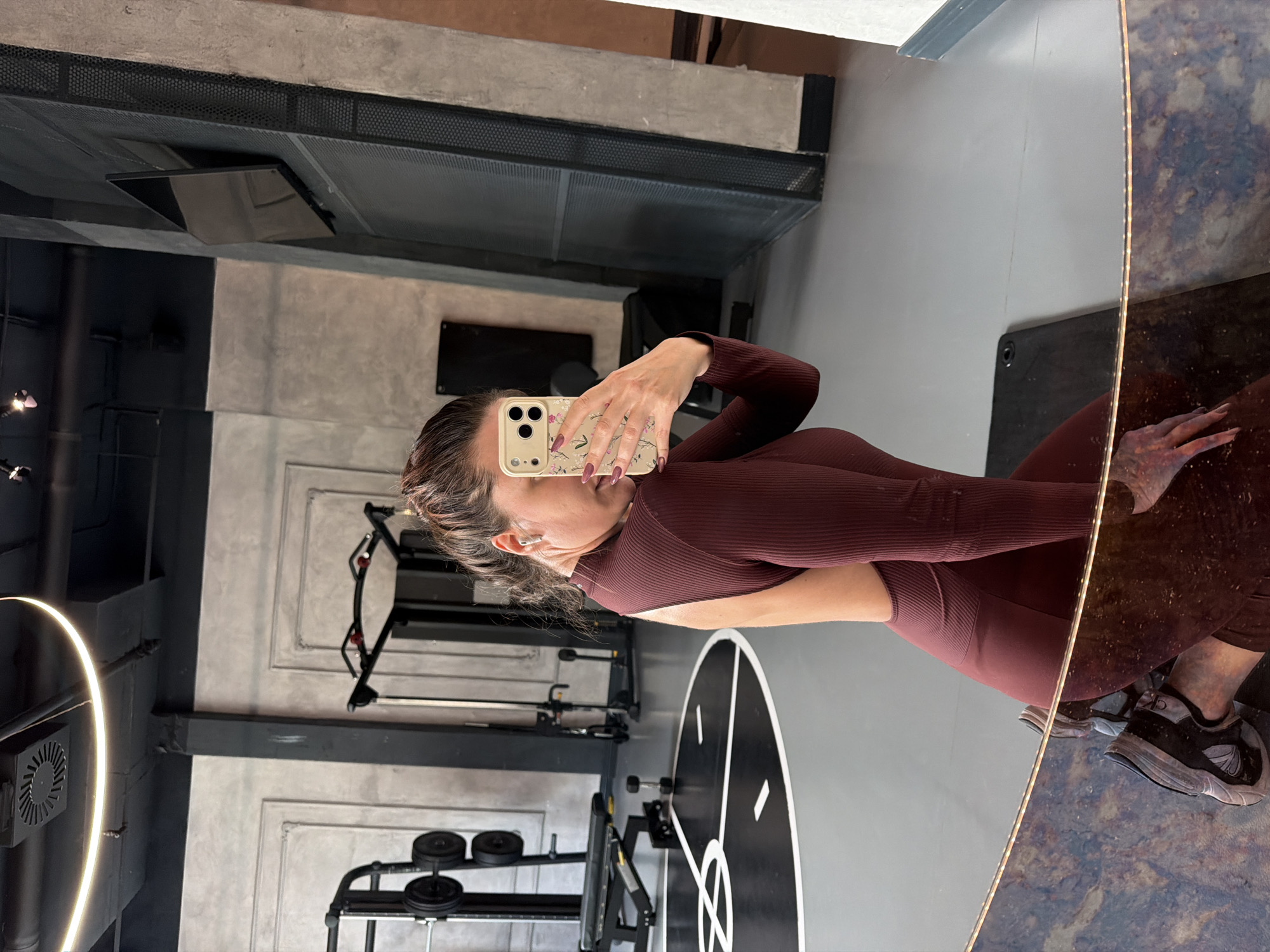 The image displays a woman taking a mirror selfie in what appears to be a gym or fitness studio. The woman, positioned on the right side of the frame, is wearing a form-fitting, ribbed burgundy long-sleeved top and matching leggings, suggesting athletic attire. Her hair is pulled back, and a white earbud is visible in her right ear. She holds a smartphone with a light-colored, floral-patterned case up to her face, obscuring most of it. Her fingernails are painted a dark color.
The setting is an indoor facility with a modern, industrial aesthetic. The background features concrete walls, some with visible paneling or texture, and an exposed ceiling structure with dark metal grates. Various pieces of gym equipment, including weight racks with dumbbells and other exercise machines, are visible in the reflections within the mirror and in the immediate surroundings. The floor is light gray with a distinct circular design in black and white, comprising geometric segments. A bright, circular light fixture is partially visible on the left. On her footwear, a "CK" logo (Calvin Klein) is faintly visible on the side of one shoe. The activity is clearly a self-portrait taken during or after a workout session at this facility, located in Belgrade, Serbia. The time of day is not determinable from the indoor lighting.