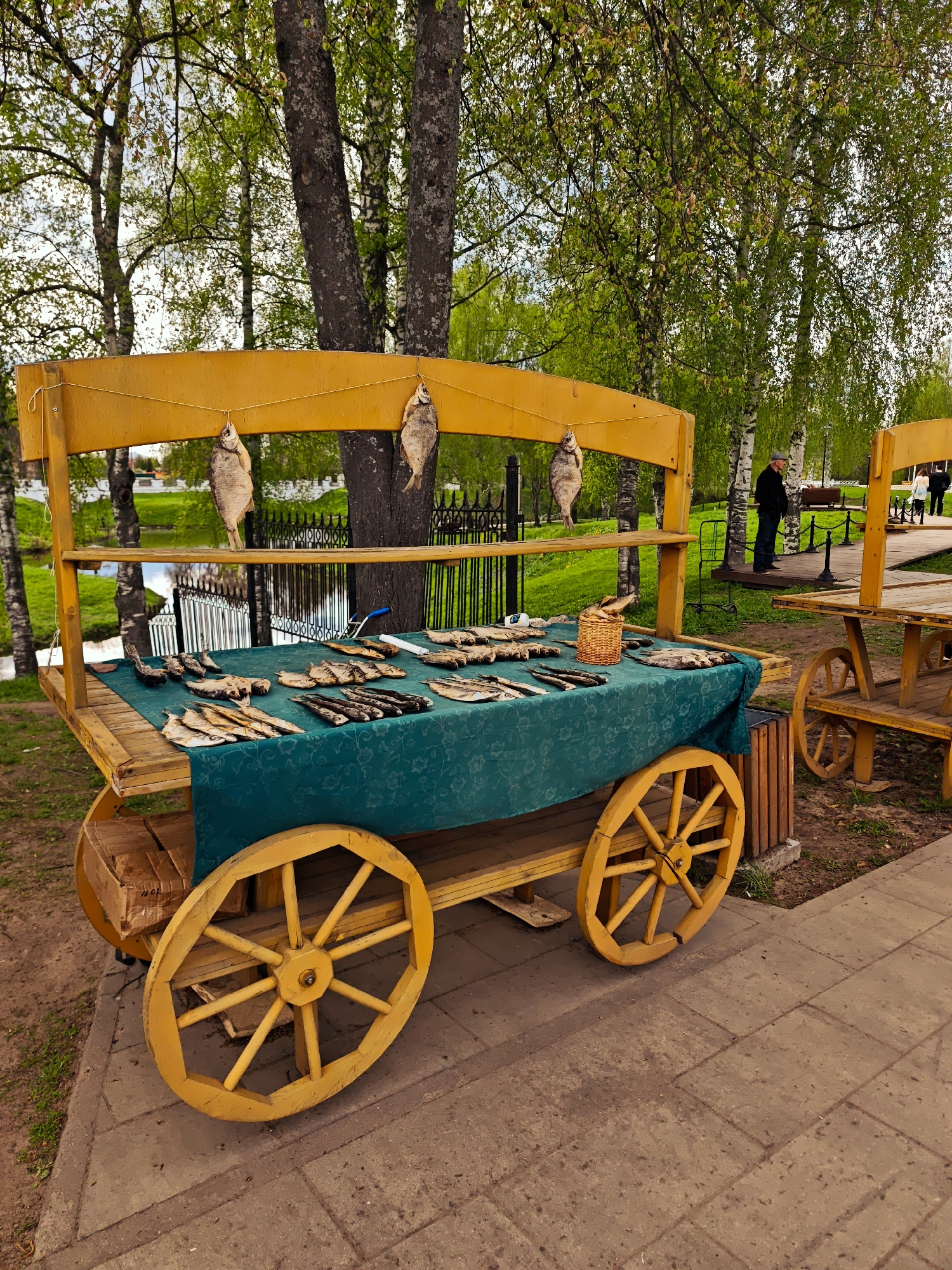 The image shows a yellow wooden cart used as a fish stand. The cart has large wheels and a green cloth covering the table-like surface. Dried fish are displayed on the cloth, and some are also hung from the upper part of the cart. In the background, there's a park-like area with trees, a pond, and a black metal fence. People can be seen walking in the distance. The location is Uglich, Russia.