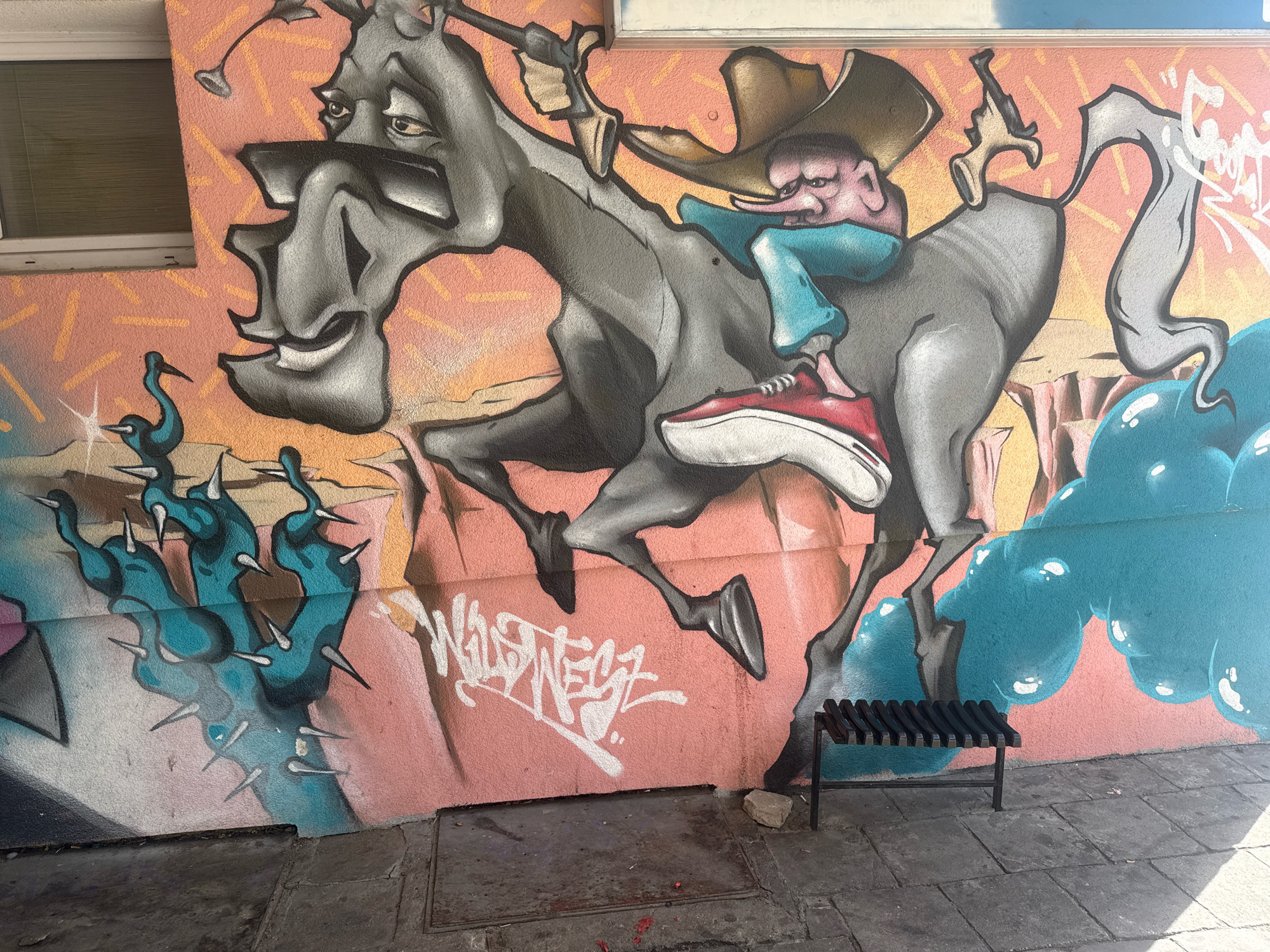The image shows a section of a mural on a textured, peach-colored building wall in Gorzow, Poland. The mural is in a graffiti style, depicting a cartoonish, anthropomorphic horse with a distorted face, ridden by a small, pink-skinned figure with a long nose wearing a cowboy hat and blue clothing. The horse is gray with black accents and has red sneakers on its hooves. To the left of the horse are several stylized, teal-colored bird-like creatures with spiky appendages. To the right, there is a large, bulbous, teal shape resembling a cloud or a cluster of balloons. A black metal bench sits on the paved sidewalk in front of the mural, with a small rock beneath it. The building has a window on the upper left side. The ground is paved with rectangular stones.