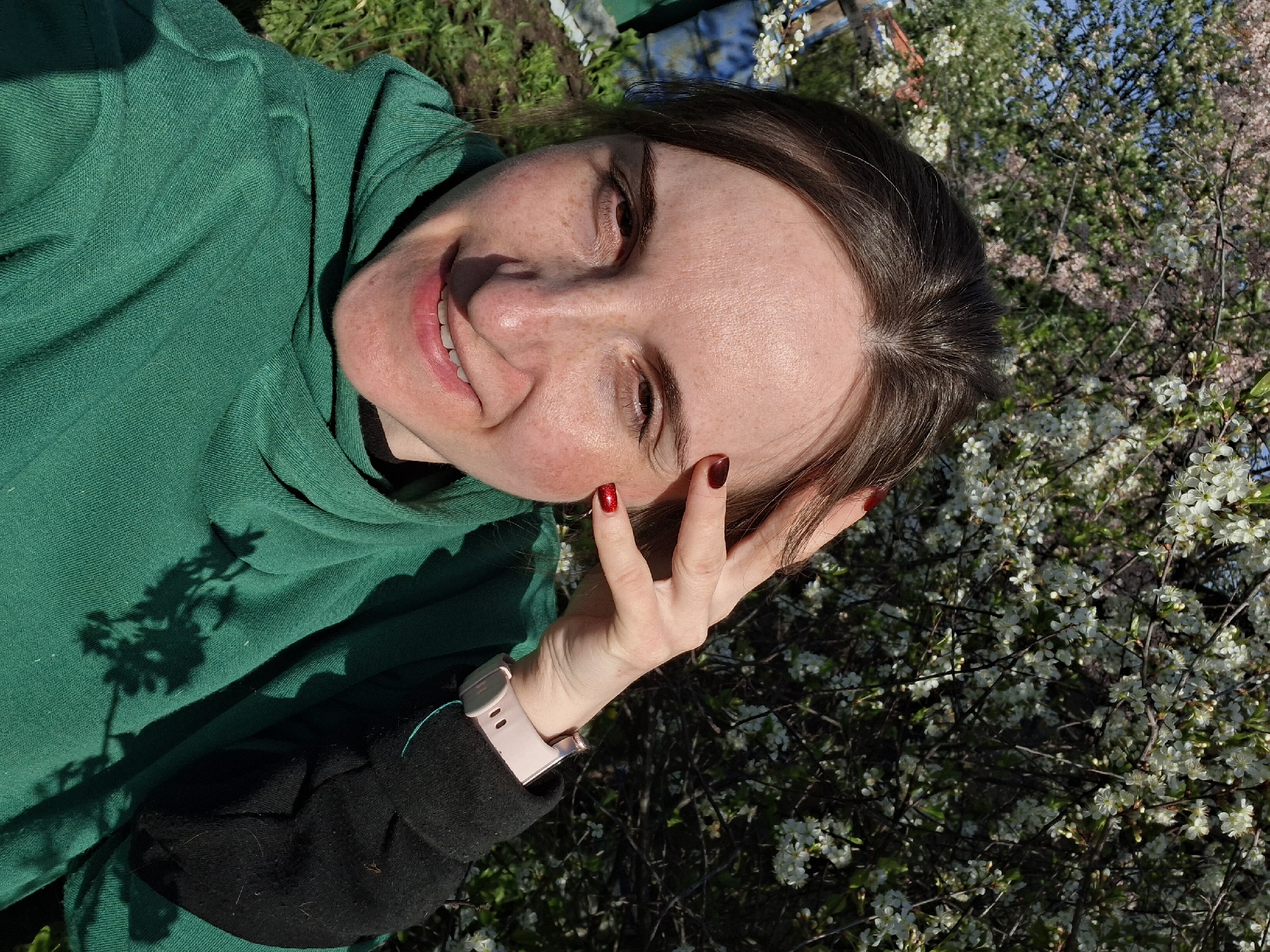 A woman is pictured in a tilted close-up shot, positioned outdoors amidst foliage and flowering trees. She wears a green hooded top, with a black sleeve portion visible on her left arm, and a pink smartwatch on her wrist. Her left hand is raised near her forehead, displaying nails painted with red polish. The background features a mix of white and pink blossoms on trees. The location appears to be a garden setting within an unspecified city, given the mix of cultivated and natural elements. The subject is looking at the camera and smiling.