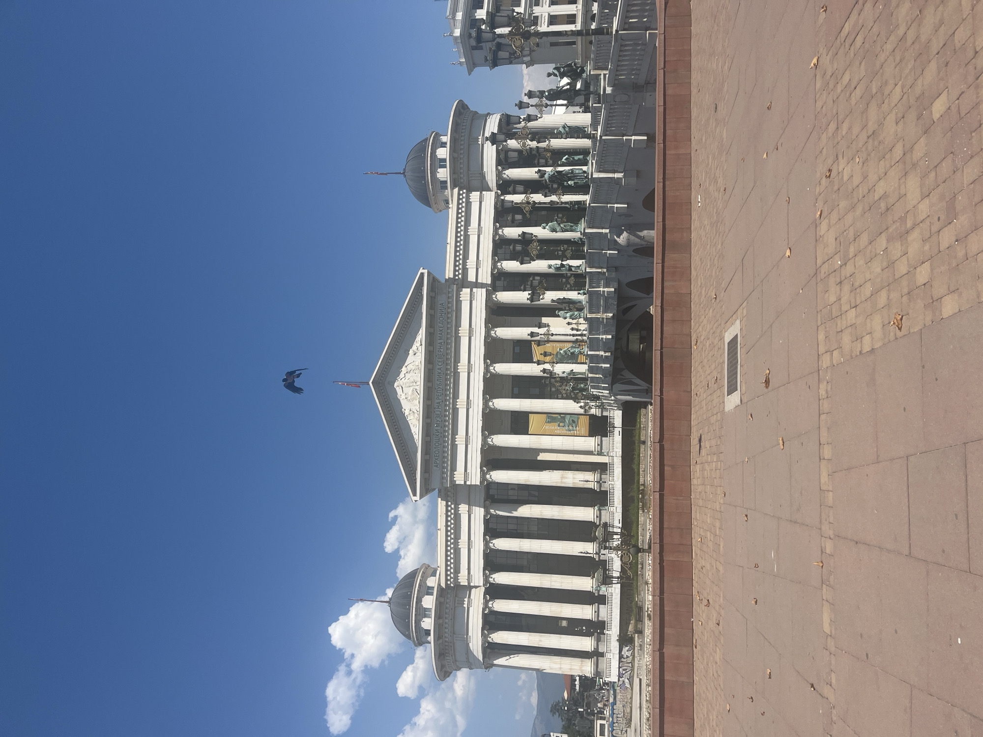A wide-angle shot captures a neoclassical building with a large portico and a dome against a clear blue sky with scattered clouds. In front of the building, a bridge lined with statues and lamps spans over a body of water. The foreground features a paved plaza with scattered dry leaves. A bird in flight is visible in the sky above the building. The building appears to be the Archaeological Museum of North Macedonia in Skopje, North Macedonia.
