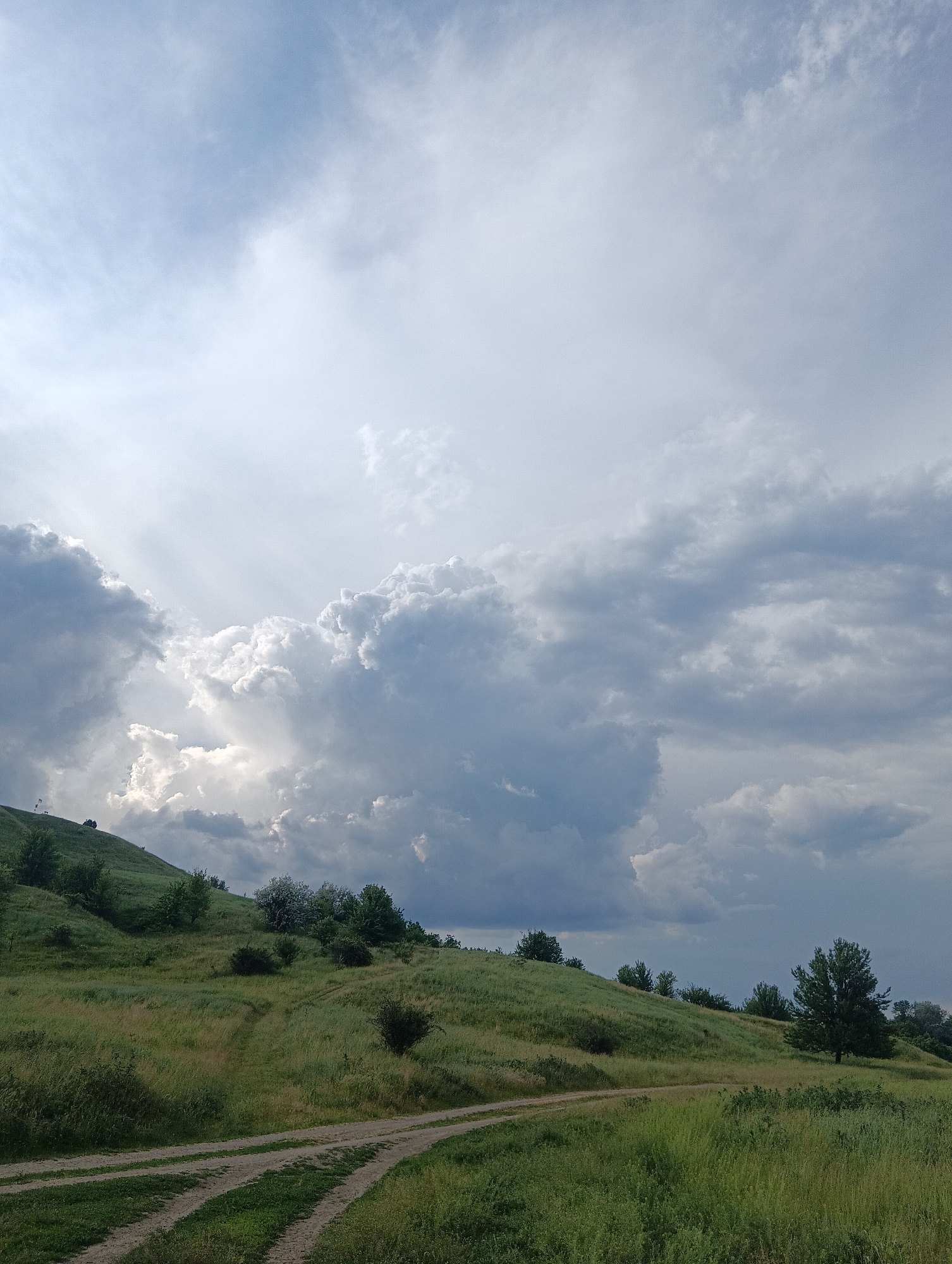 The image displays a hilly, rural landscape under a dynamic sky. The upper two-thirds of the frame are occupied by a complex cloud system, featuring bright white, sun-illuminated cumulus and cumulonimbus clouds predominantly on the left and central portions, where distinct crepuscular rays are visible. These bright clouds contrast with darker grey cloud masses, particularly in the upper right and lower central sky. The lower third of the image depicts gently rolling green hills covered in lush grass and interspersed with various deciduous trees and bushes. A light-brown dirt track, comprising multiple parallel paths, curves from the bottom-center-left into the mid-ground. On the crest of the leftmost hill, a faint white structure, possibly a small telecommunications mast or observation post, with an attached white panel and a small flag, is visible. No human figures or discernible animals are present. The scene is located in Unknown, Unknown.