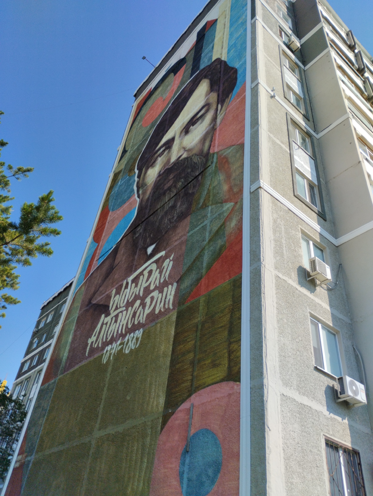 The image shows a large mural painted on the side of a multi-story building, likely an apartment complex, in Kostanay, Kazakhstan. The mural depicts a portrait of a man with a beard and a hat, rendered in a stylized, colorful manner with abstract geometric shapes in the background. The man's name, "Ыбырай Алтынсарин" (Ibyrai Altynsarin), and the dates "1841-1889" are visible in white lettering on the mural. The building itself is made of concrete with visible windows and air conditioning units. Part of a pine tree is visible in the foreground on the left side of the image, and the sky is a clear blue. The perspective is from a low angle, looking up at the building.