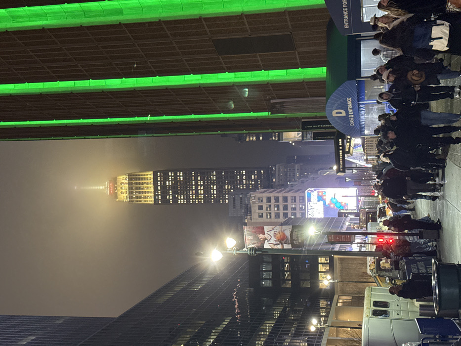 The Empire State Building rises through fog at night, lit with yellow windows and a red and yellow top. Green horizontal lights line the bottom of an elevated structure above. Below, a crowd of people lines up near a "Chase Entrance" sign. Streetlights illuminate the foreground, and city buildings flank the sides. A basketball poster is visible amidst the city lights.