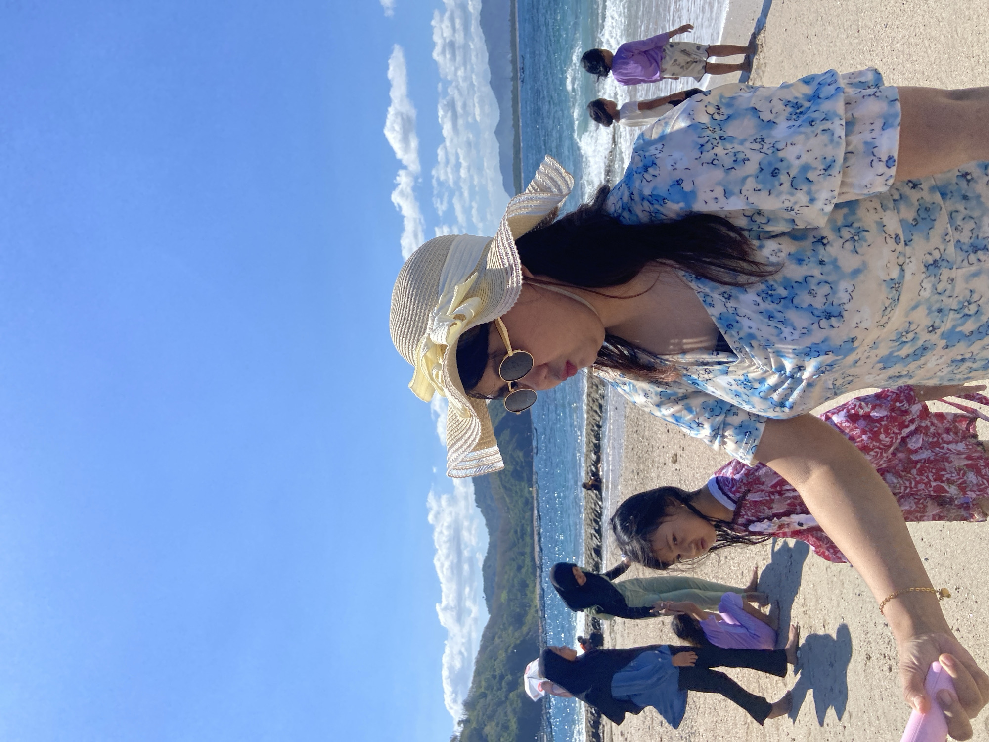 A bright outdoor shot captures a group of people on a beach, likely in Unknown, Indonesia. A woman in a blue floral patterned dress, a light-colored sun hat, and round sunglasses is prominently featured in the foreground, positioned at an angle. Beside her, a young girl in a red patterned dress is also looking down. The woman is holding a pink object.
Further back on the beach, other individuals are visible. A woman dressed in black stands near the shoreline, and another figure in a purple shirt stands closer to the water's edge, interacting with the waves.
The background reveals a coastline with mountains and a clear blue sky with scattered clouds. The ocean is turquoise near the beach and deep blue further out. A low wall or rock formation extends into the water, separating the sandy beach from the sea.
