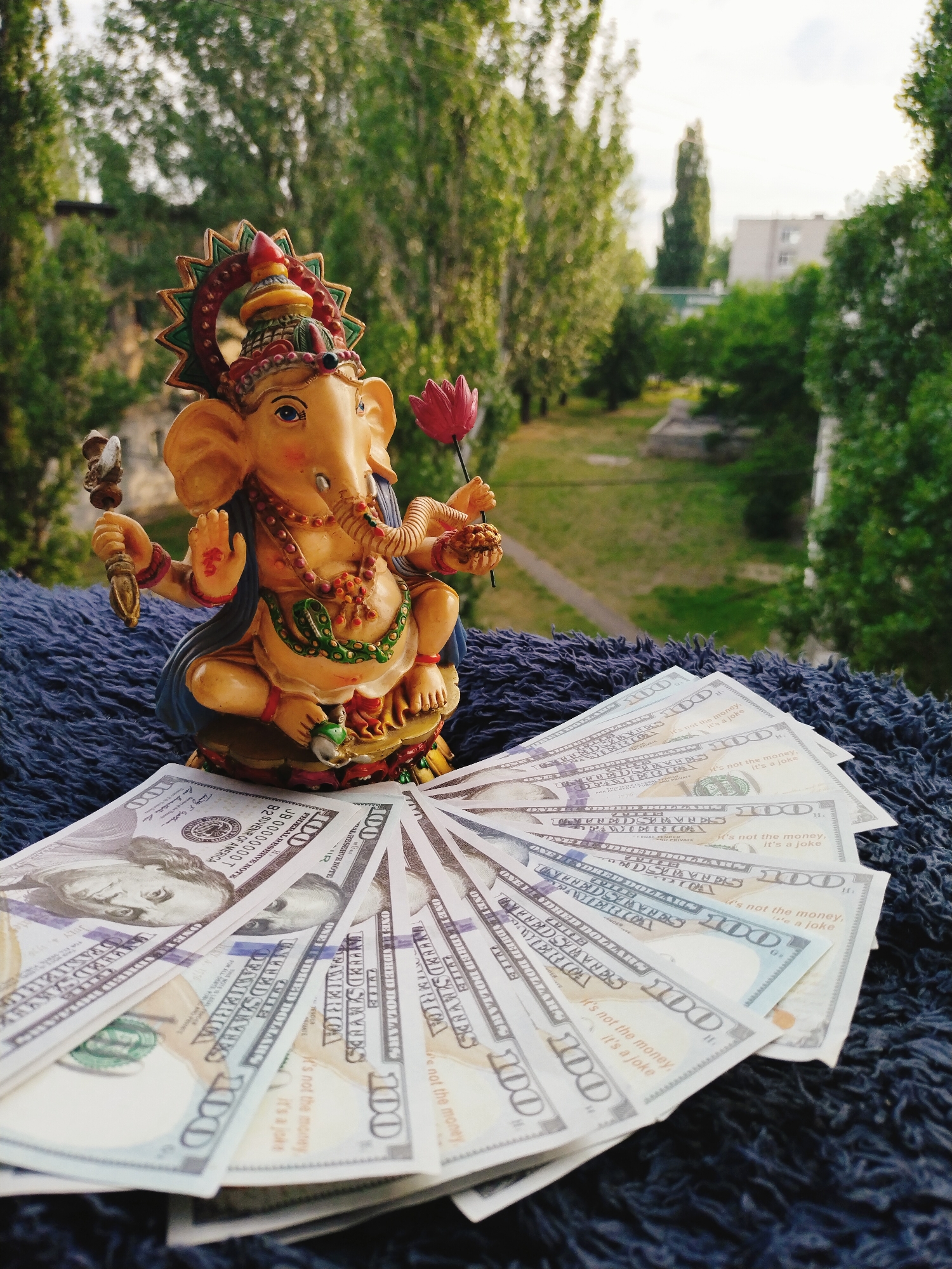 A small, colorful statue of the Hindu deity Ganesha, an elephant-headed god, sits on a dark blue, textured surface. Ganesha is depicted with multiple arms, holding various objects, including a lotus flower. In front of the statue, a fan of US hundred-dollar bills is spread out. The background shows a blurred view of trees and buildings, suggesting an outdoor setting, possibly a balcony or windowsill, overlooking a cityscape. The scene is illuminated by natural light, creating a soft, even exposure. There are no people present in the image.